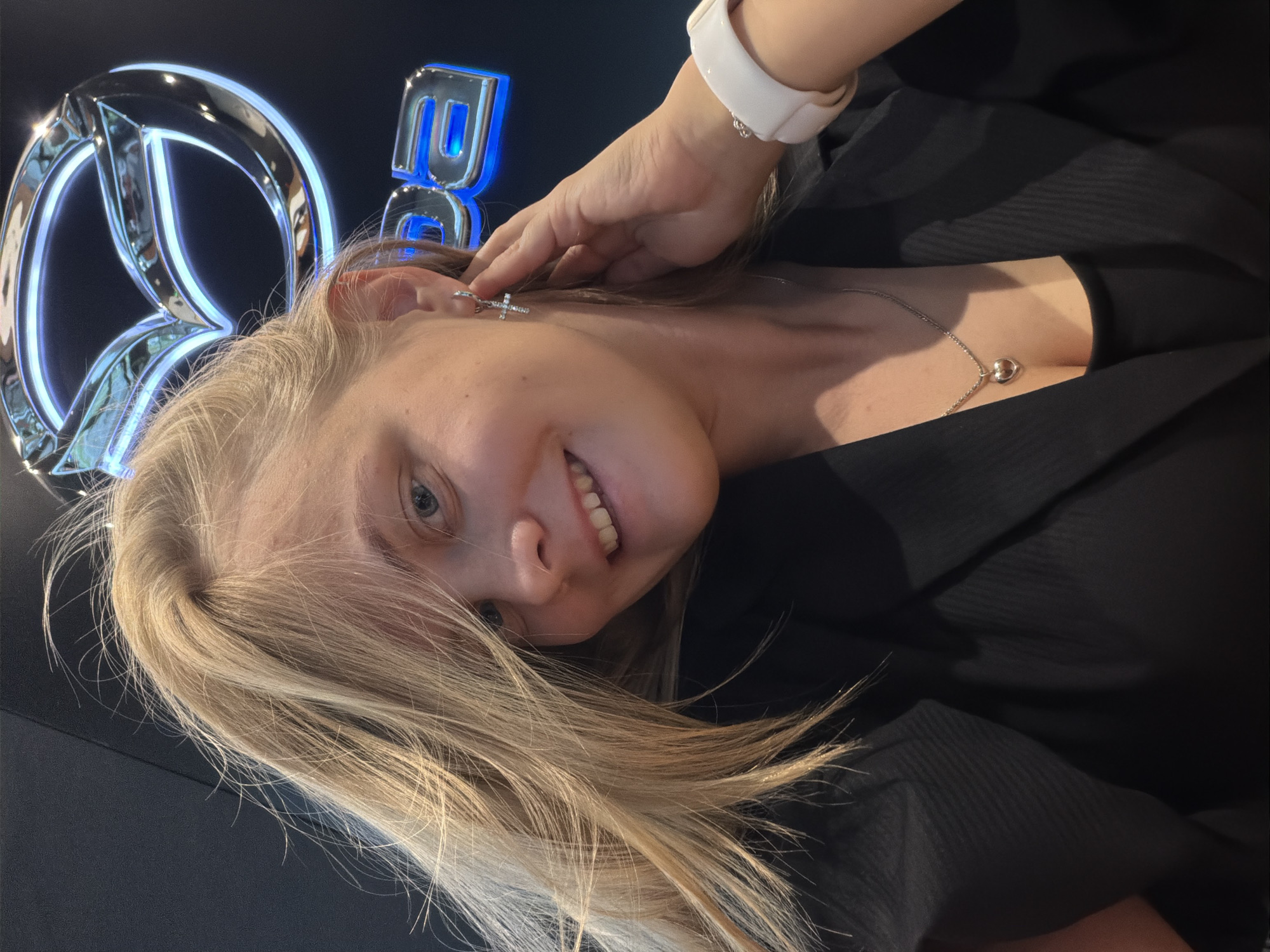 A woman with light blonde hair is looking at the camera and smiling. She is wearing a black top, a white watch and a silver necklace. She is standing in front of a brightly lit Mercedes-Benz logo. A hand is touching her hair near the logo. It is highly likely that this photo was taken in Izhevsk, Russia, at an event related to Mercedes-Benz automobiles.