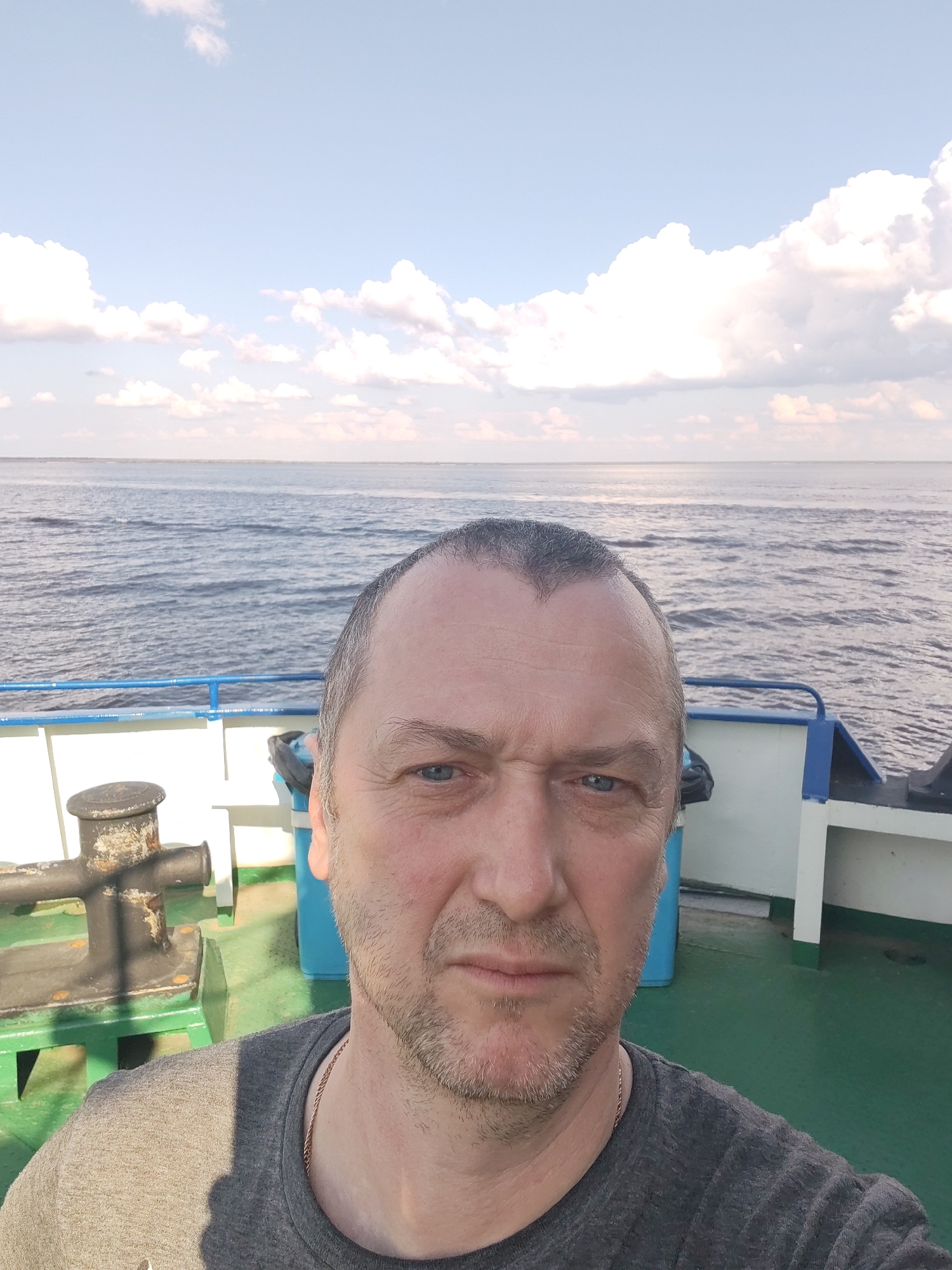 A middle-aged man takes a selfie onboard a vessel, likely a boat or ferry, near the city of Veregonets, Russia. He has short, graying hair, blue eyes, and light stubble. The background shows a body of water meeting the horizon under a partly cloudy sky. The vessel has a green deck with a black mooring bollard, white and blue railings, and what appears to be blue recycling bins. The man is wearing a gray t-shirt and a gold chain around his neck.