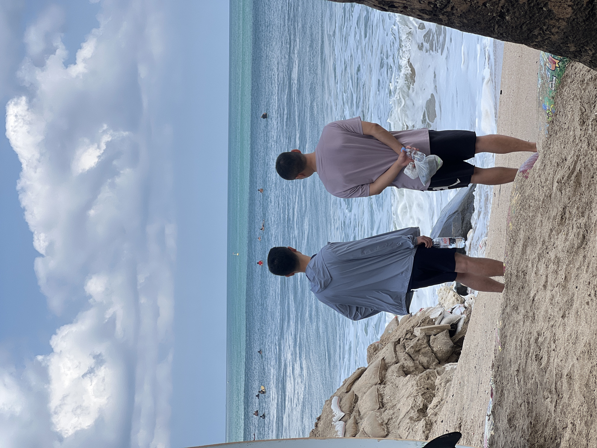 Two individuals stand on a sandy beach, facing the ocean in Kuta, Indonesia. The sky is cloudy, and the ocean is turquoise with white-capped waves. Several surfers are visible in the water. The beach is littered with debris, including sandbags forming a makeshift barrier on the left and a fallen tree trunk on the right. One person is wearing a gray jacket and black shorts, holding a bottle of water. The other person is wearing a pink t-shirt and black shorts, with their hands clasped behind their back, holding a plastic bag and a bottle of water. A surfboard is partially visible on the far left.