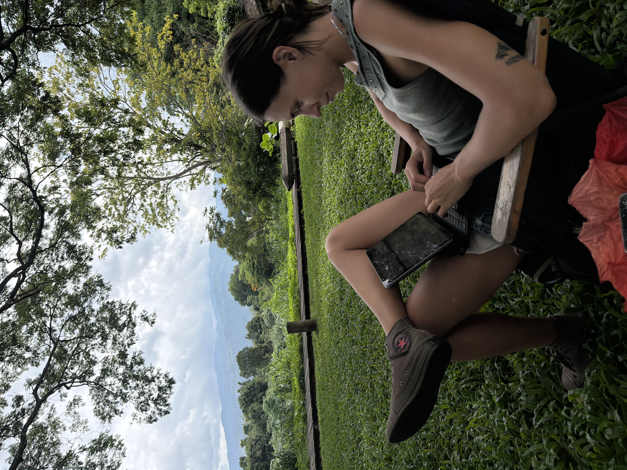 A woman is seated outdoors in a chair, looking down at a tablet device. She is wearing a sleeveless olive green top and dark shorts, with brown high-top Converse sneakers. Her legs are crossed, with one foot resting on a low wooden railing. The tablet is positioned on her lap, and her hands are on the keyboard. She has dark hair pulled back in a ponytail and a silver necklace. In the background, there is a lush green lawn, a wooden pathway, and dense foliage. Beyond that, a range of mountains is visible under a cloudy sky. The setting appears to be a natural, possibly tropical, environment.