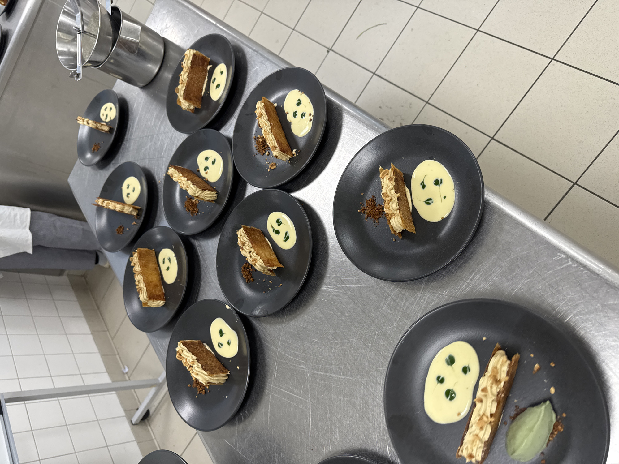 The image shows several plates of desserts arranged on a stainless steel countertop in what appears to be a commercial kitchen setting. The plates are dark grey and each contains a rectangular pastry with cream filling, a dollop of light-colored sauce with green dots, and some crumbs or small pieces of garnish. In the upper left corner, there's a stainless steel container with some utensils. The background includes white tiled walls and a stainless steel table. The location is Schiltigheim, France.