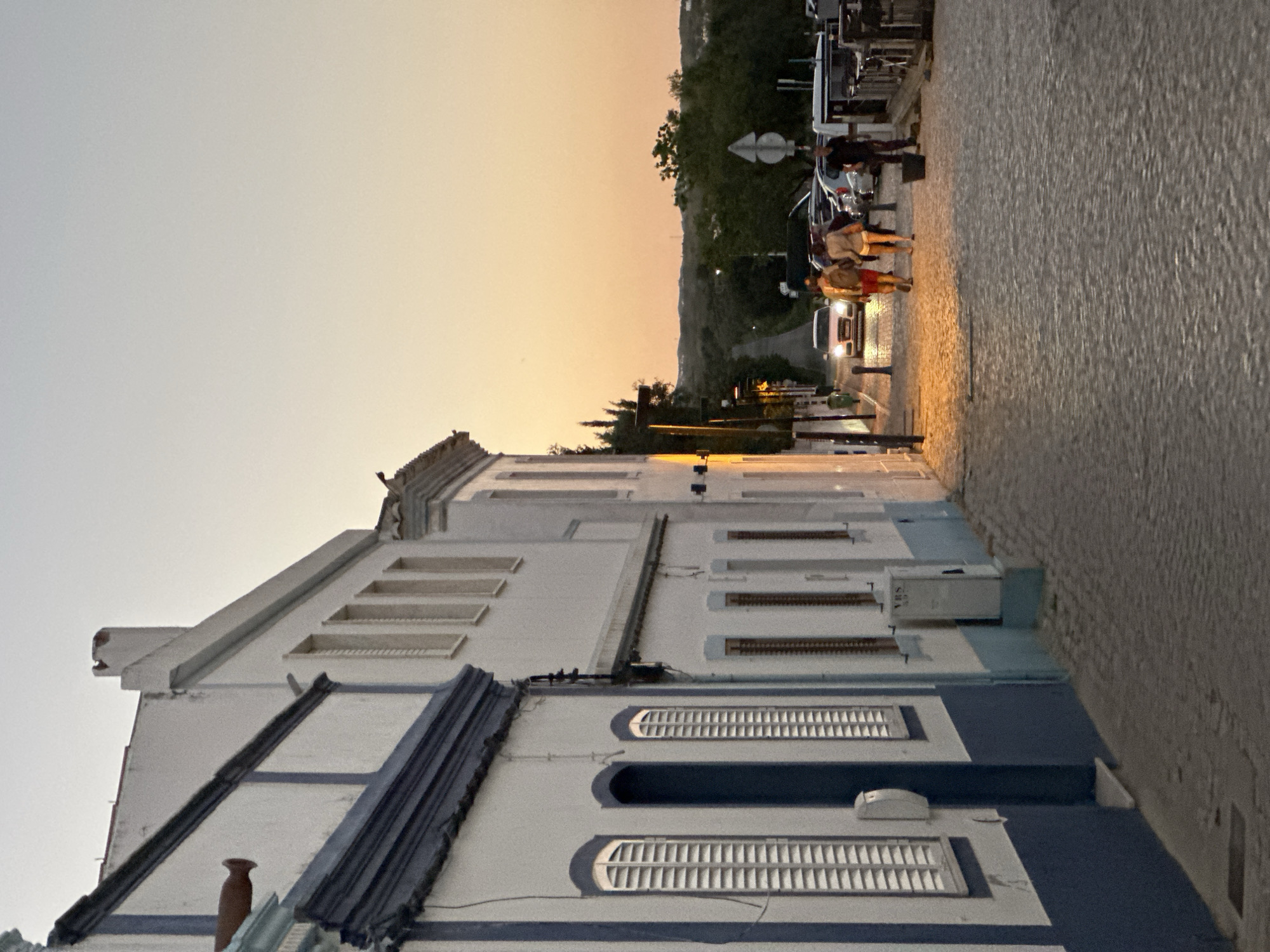 This image captures a view from a street in Vila Real de Santo António, Portugal, at dusk or dawn. The foreground shows a row of white buildings with blue trim, a common architectural style in the region. The buildings have multiple windows with shutters.
On the right side of the frame is a cobbled street, where several people are walking. Further along the street, cars are parked, and some are moving, their headlights visible. The background transitions into a hazy sky with a gradient of yellow and orange hues, suggesting either sunrise or sunset. Some trees are visible in the background.