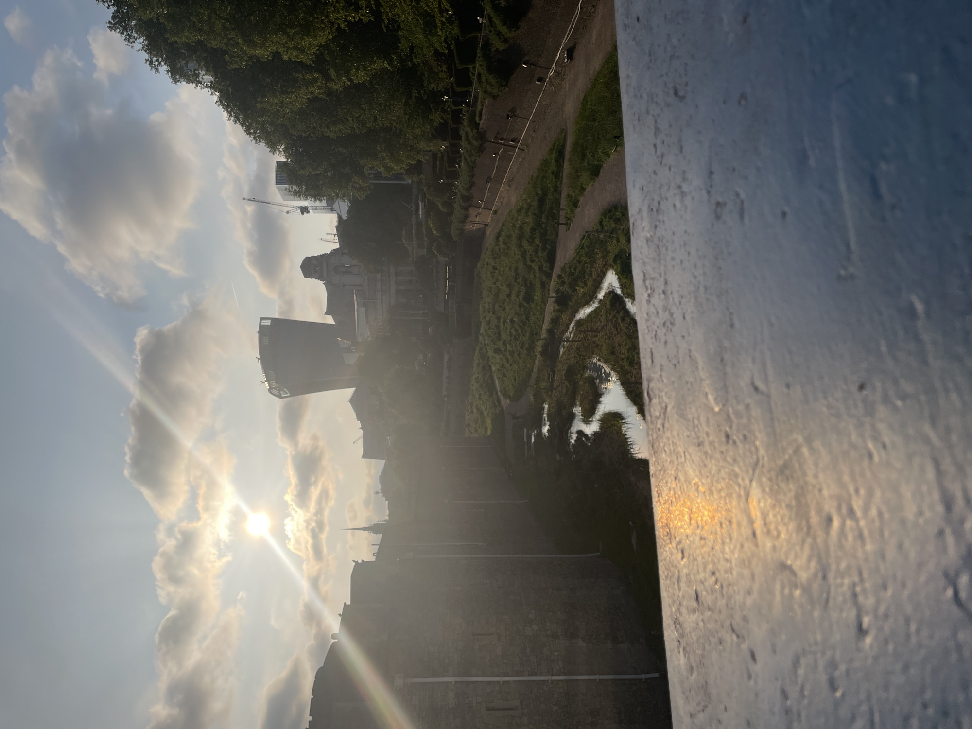 The image captures a view in London, United Kingdom, looking towards the Tower of London complex. The sun is prominent, partially obscured by clouds, casting a bright glare across the scene. In the foreground, a white painted structure, possibly a bridge railing, takes up a significant portion of the right side. Beyond this, a landscaped green area with pathways and a small body of water is visible. Further in the background, the distinct stone walls and buildings of the Tower of London are present, with a modern tall building rising behind them. A construction crane is visible amongst the city structures. A line of small evenly spaced lights runs along the path near the grassy area.