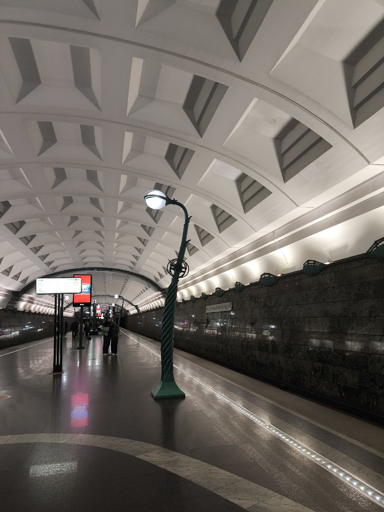 The image shows a subway station platform. The station has an arched ceiling with a repeating pattern of square indentations. A green, decorative lamppost stands prominently on the platform. Along the wall, there are mounted lighting fixtures. People are seen standing and walking on the platform. A digital display board is visible in the background. The floor is a patterned marble or stone. A line of lights is embedded in the floor, running alongside the platform edge.