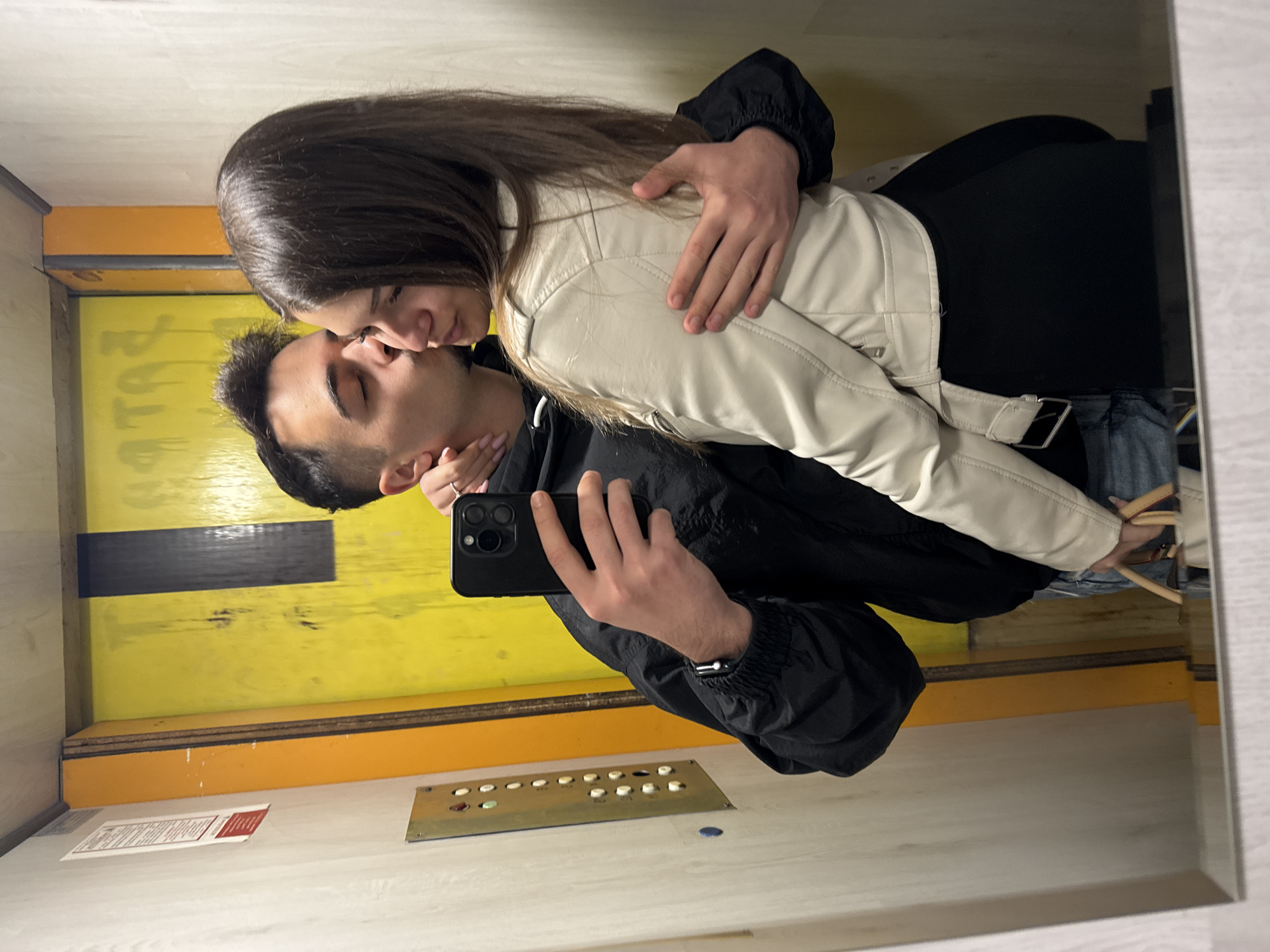 A mirror selfie captures a young couple sharing an intimate moment inside an elevator. The image is taken from the perspective of the male, who holds a black smartphone to photograph himself and the female embracing. The female, with long dark hair, wears a cream-colored leather-style jacket over a dark top and blue jeans, wrapping her left arm around the male's neck as they kiss. The male, in a black jacket, has his left arm around her waist.
The elevator interior features distinctive bright yellow walls with some darker horizontal bands, suggesting an older or utilitarian design. The floor is a reflective, light-colored surface, mirroring parts of the subjects and the elevator's features. A metal control panel with multiple circular buttons, some illuminated, is visible on the lower wall. To the left of the control panel, a white paper with red text is partially visible on the floor, bearing the Bulgarian word "ИНФОРМАЦИОНЕН" (INFORMATION). A glimpse of a light brown, possibly woven, bag or purse handle can be seen near the female's right hip. The overall atmosphere conveys a moment of affection between the couple. The setting is likely in Sofia, Bulgaria, given the visible text and location context.