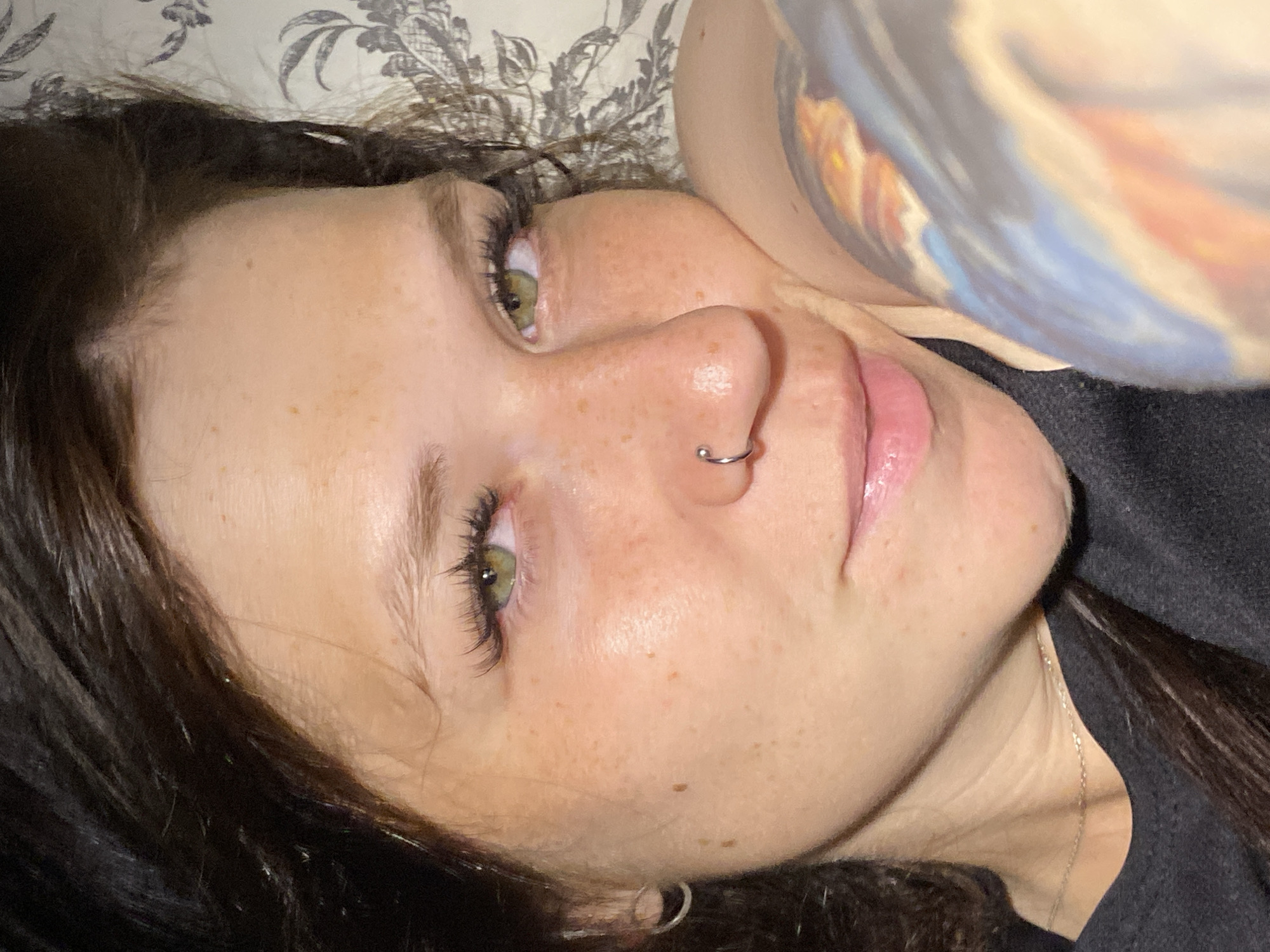 A close-up, high-angle portrait of a young woman with dark hair and green eyes, lying down with her head resting on her right arm, which is partially visible and adorned with a colorful tattoo. Her left ear, adorned with a silver hoop earring, is visible. She is wearing a dark gray crewneck shirt. Her face is partially illuminated, highlighting freckles across her cheeks and nose. She has a silver hoop nose ring. The background features a floral-patterned fabric. The image is believed to have been taken in Chaikovsky, Russia.