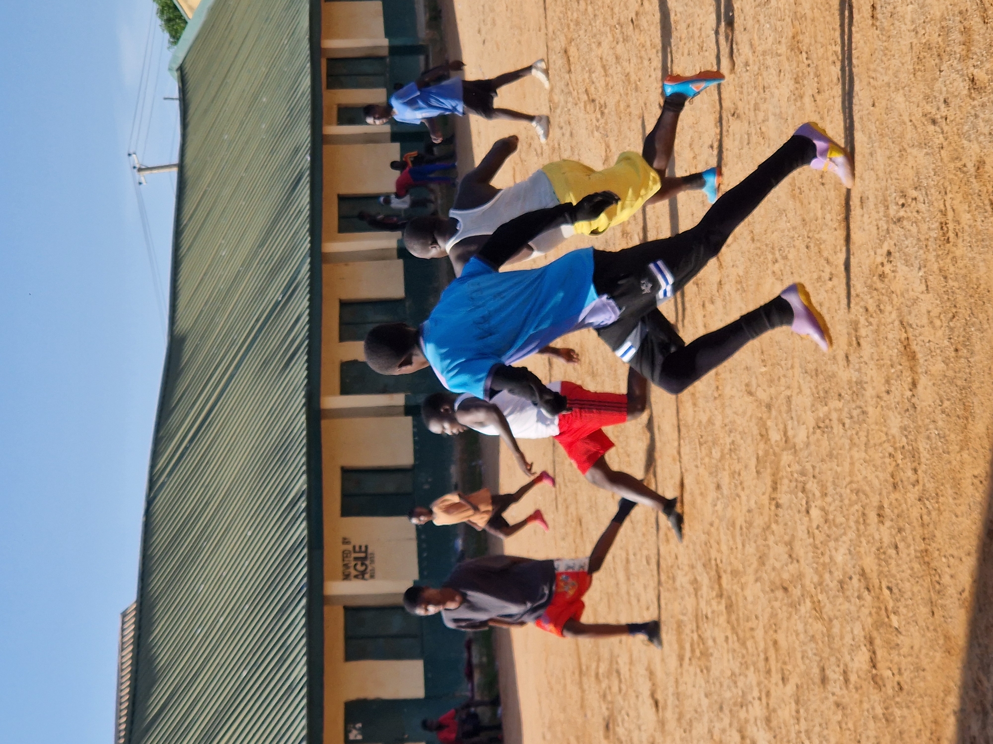 The image shows an outdoor soccer practice or game at an educational facility in Kabong, Nigeria. Several young boys are actively engaged in the sport on a dusty, orange-colored field. Some are in motion, running and kicking, while others appear to be observing or preparing to join the activity. They are wearing athletic attire, including shorts, t-shirts, and soccer cleats in various colors. In the background, a low-rise building with a green, corrugated metal roof and light-colored walls is visible. The building has multiple windows and doors. The sky is clear and blue. The overall atmosphere suggests a typical day of sports activities at a school or community center.