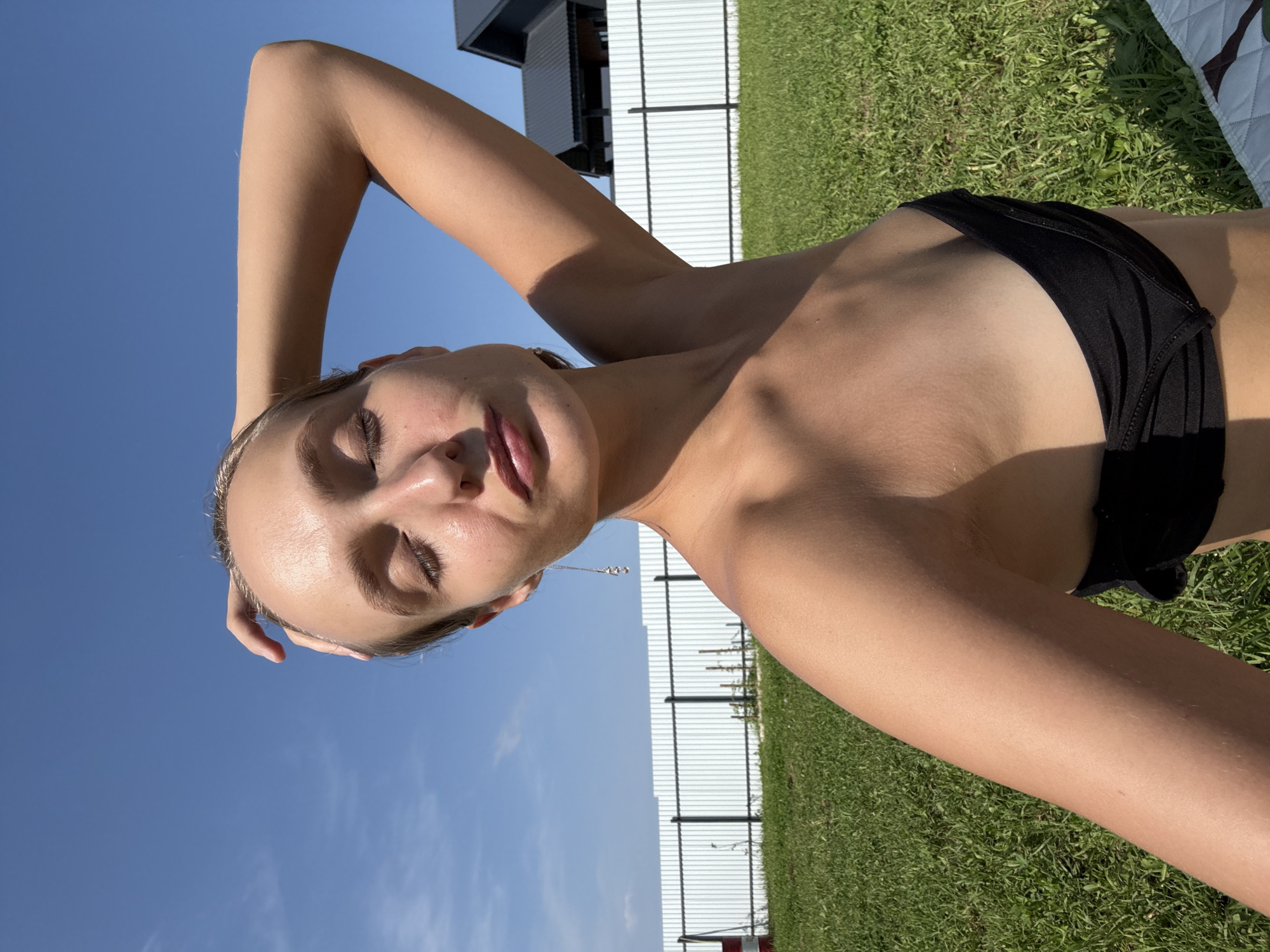 A woman is lying down on grass, with the sky visible in the background. She is wearing a black bikini top and has her arm behind her head. Her eyes are closed and her lips are slightly parted. The sun is shining brightly, casting shadows on the grass. In the background, there is a white fence and a modern building. The scene appears to be set in a backyard or park on a sunny day. The woman's pose and relaxed expression suggest she is enjoying the warm weather and possibly sunbathing. The overall mood is serene and peaceful. There is no visible text in the image. The location context provided is Подшивалово, Russia.