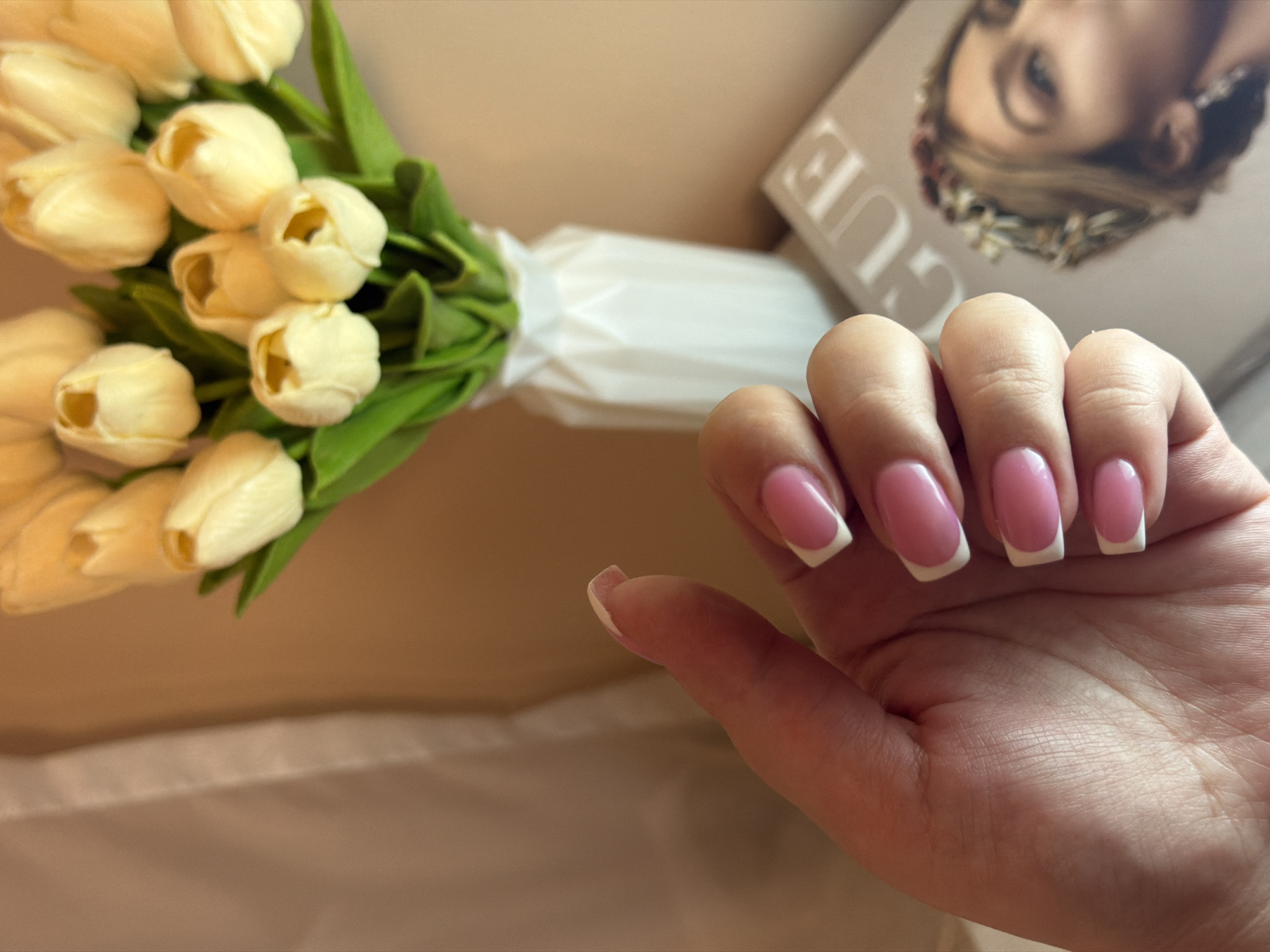 A hand with pink and white French manicure nails is positioned in the foreground of the image. Behind the hand to the left, there is a bouquet of cream-colored tulips wrapped in white paper. To the upper right behind the hand, there is a copy of Vogue magazine. The background appears to be a beige fabric surface.