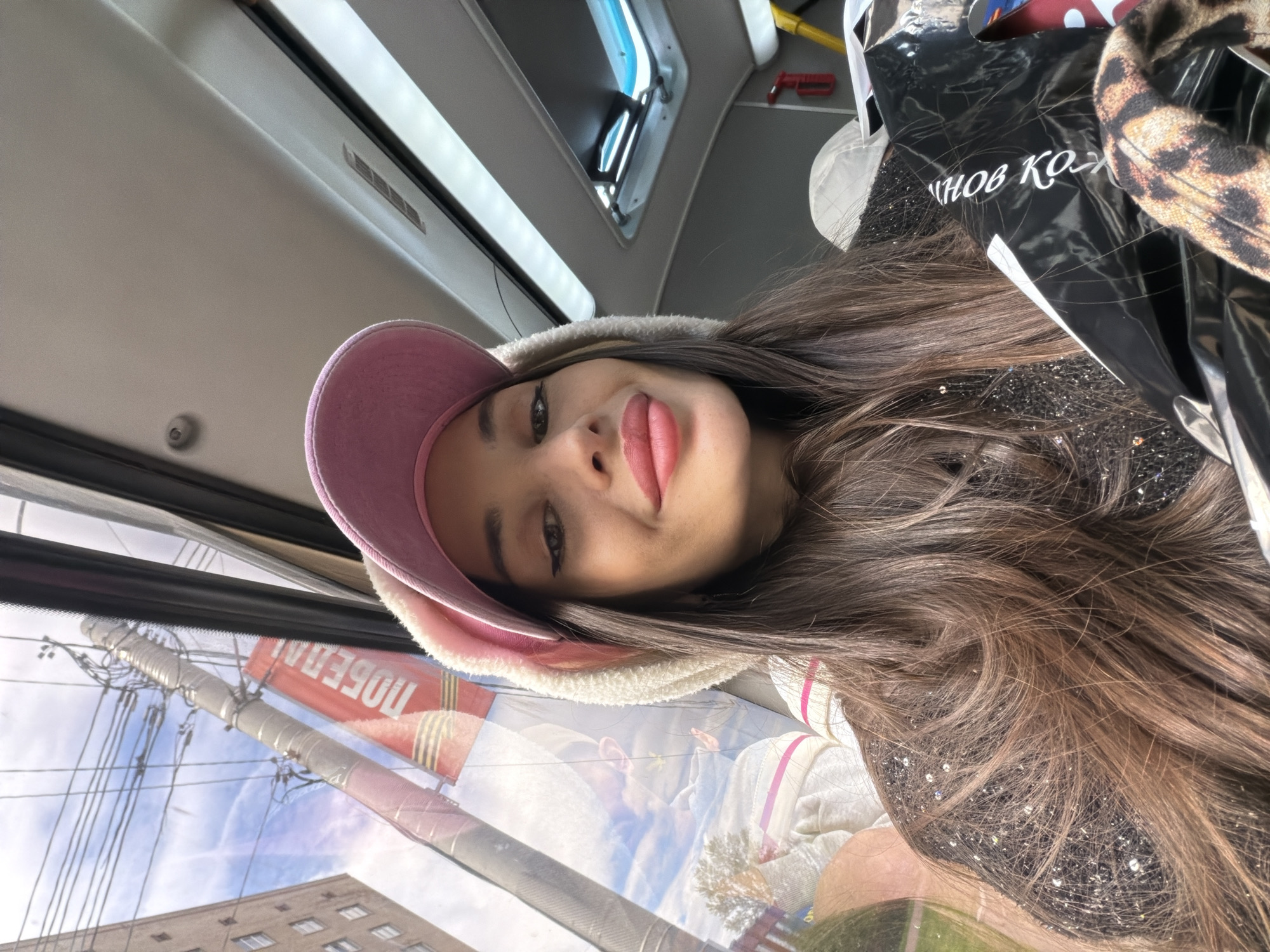 A young woman wearing a pink baseball cap with faux fur trim is inside a public bus. She is looking at the camera. She is holding a black shopping bag. Through the bus window, a power pole and lines with a red banner that says "Победа" can be seen. This indicates the image was taken in Saint Petersburg, Russia.