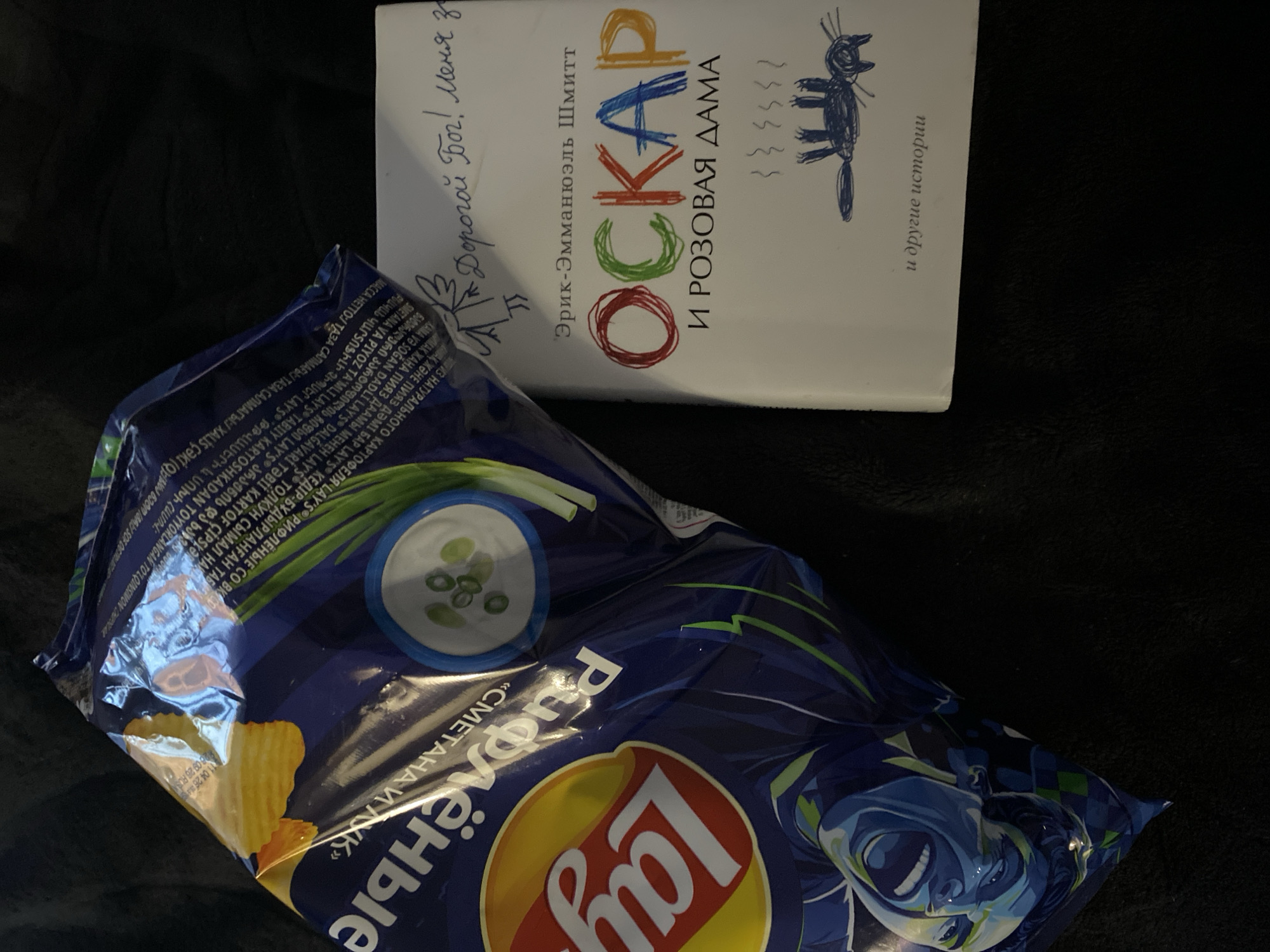 The image shows a bag of Lay's "Cream & Onion" ridged potato chips next to the Russian edition of the book "Oscar and the Lady in Pink" by Éric-Emmanuel Schmitt. The book has a handwritten note on the cover that says, "Dear God! Меня з...", which translates to "Dear God! Me...". The scene appears to be indoors on a dark surface. The image does not provide enough information to determine the location in Saint Petersburg or any specific interactions taking place.