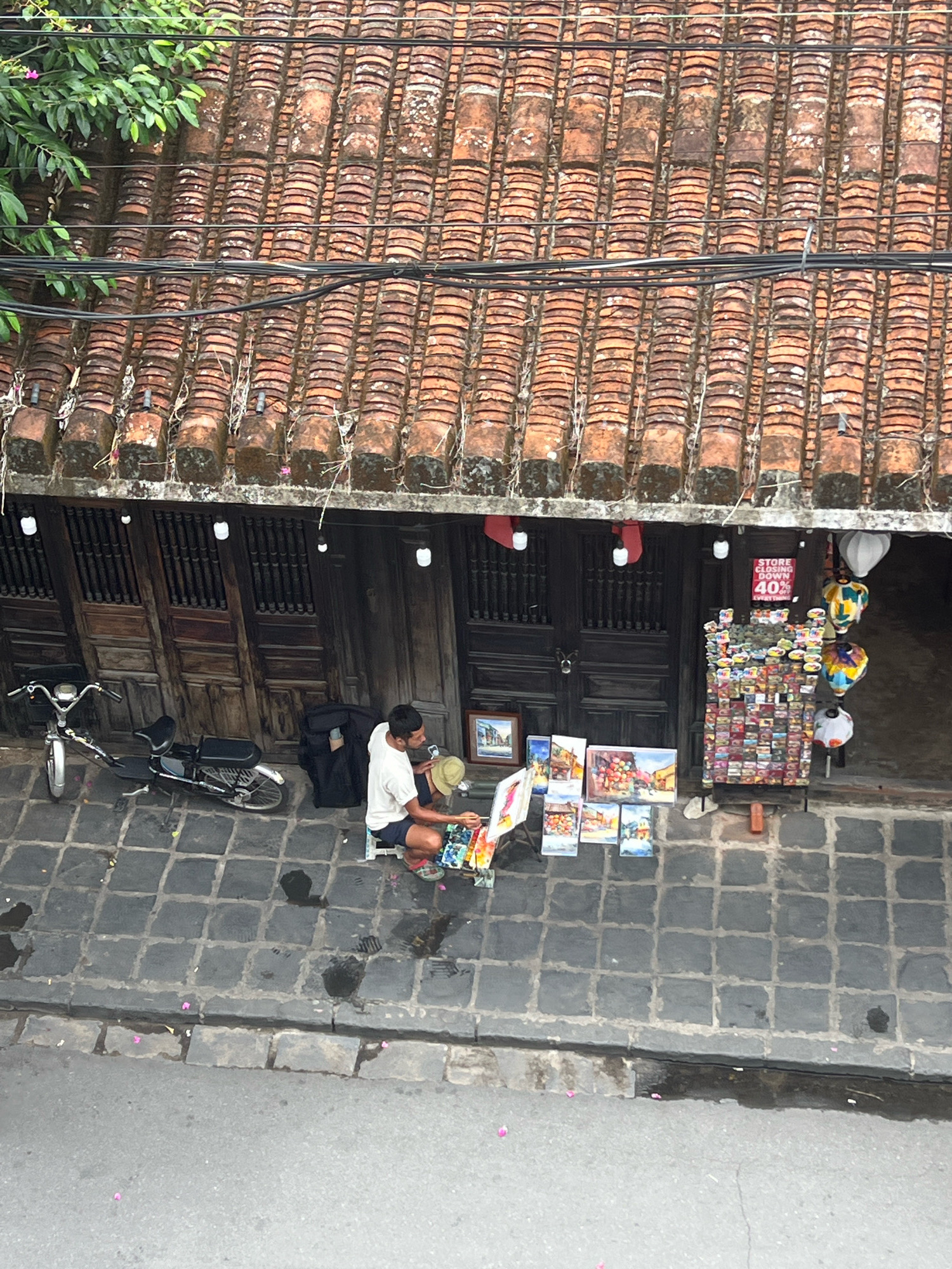 The image captures an overhead view of a street scene in Hoi An, Vietnam. The foreground shows a wet asphalt road with scattered pink petals. A grey tiled gutter separates the road from the sidewalk, which is paved with dark grey square tiles. On the sidewalk, a man is seated on a low stool, facing a display of paintings and other items. He is wearing a white t-shirt and dark shorts, and a straw hat rests near him. To his left, a black electric scooter is parked. Behind the man, a shop with traditional wooden doors is visible. A display rack filled with small colorful items, possibly souvenirs or artwork, stands against the shop. Several lanterns, some patterned and some white, hang near the shop entrance. The shop's roof is covered with weathered, orange clay tiles. The overall scene depicts a commercial street in Hoi An, likely in the historic ancient town, with a vendor selling artwork.