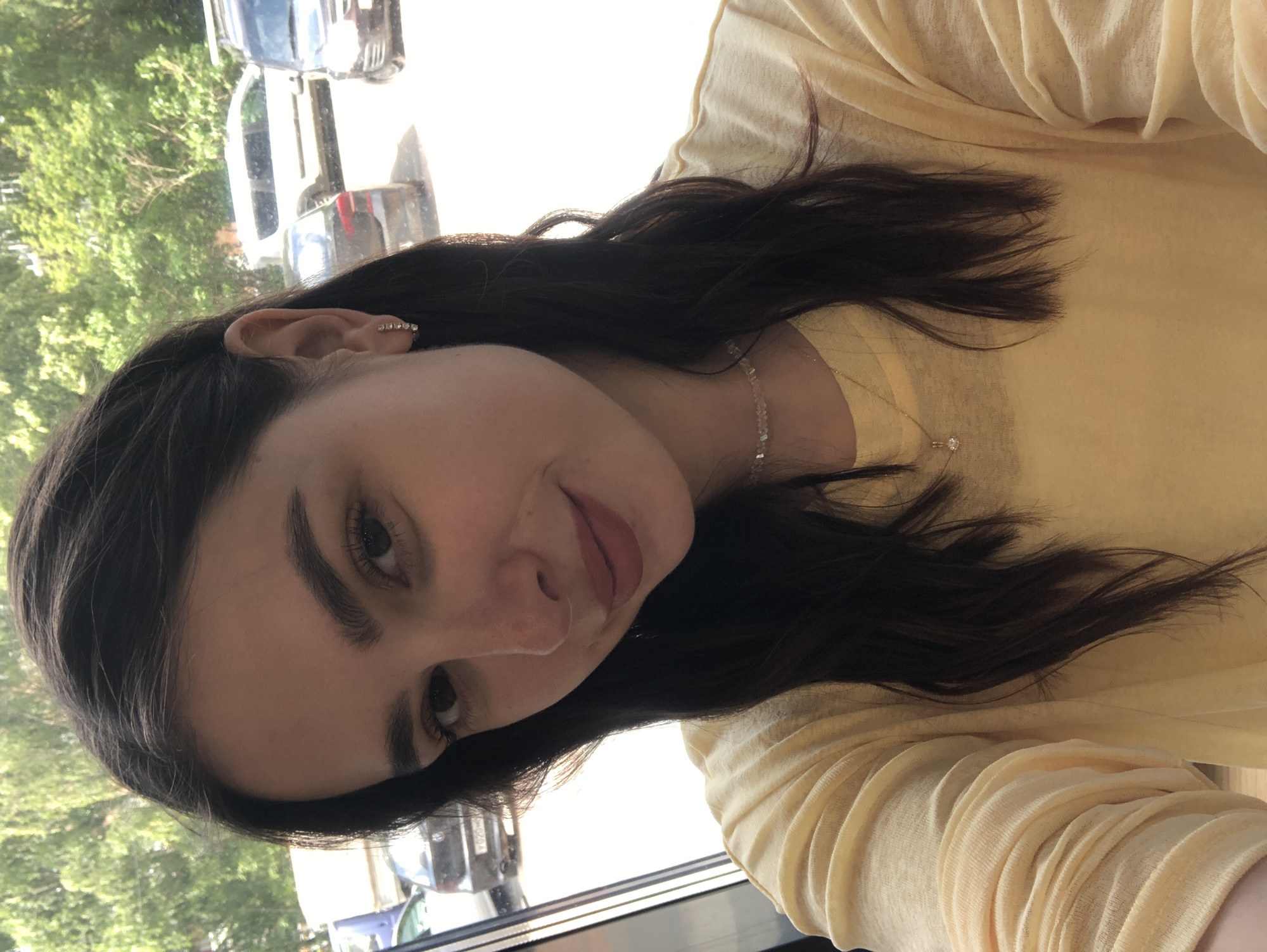A young woman with dark hair is taking a selfie from a low angle. She is wearing a yellow t-shirt and a delicate necklace. Her hair is dark brown and falls around her shoulders. She has dark eyebrows and dark eyes. The background appears to be a street scene with trees and cars visible through what looks like a window. The overall lighting suggests it is daytime.