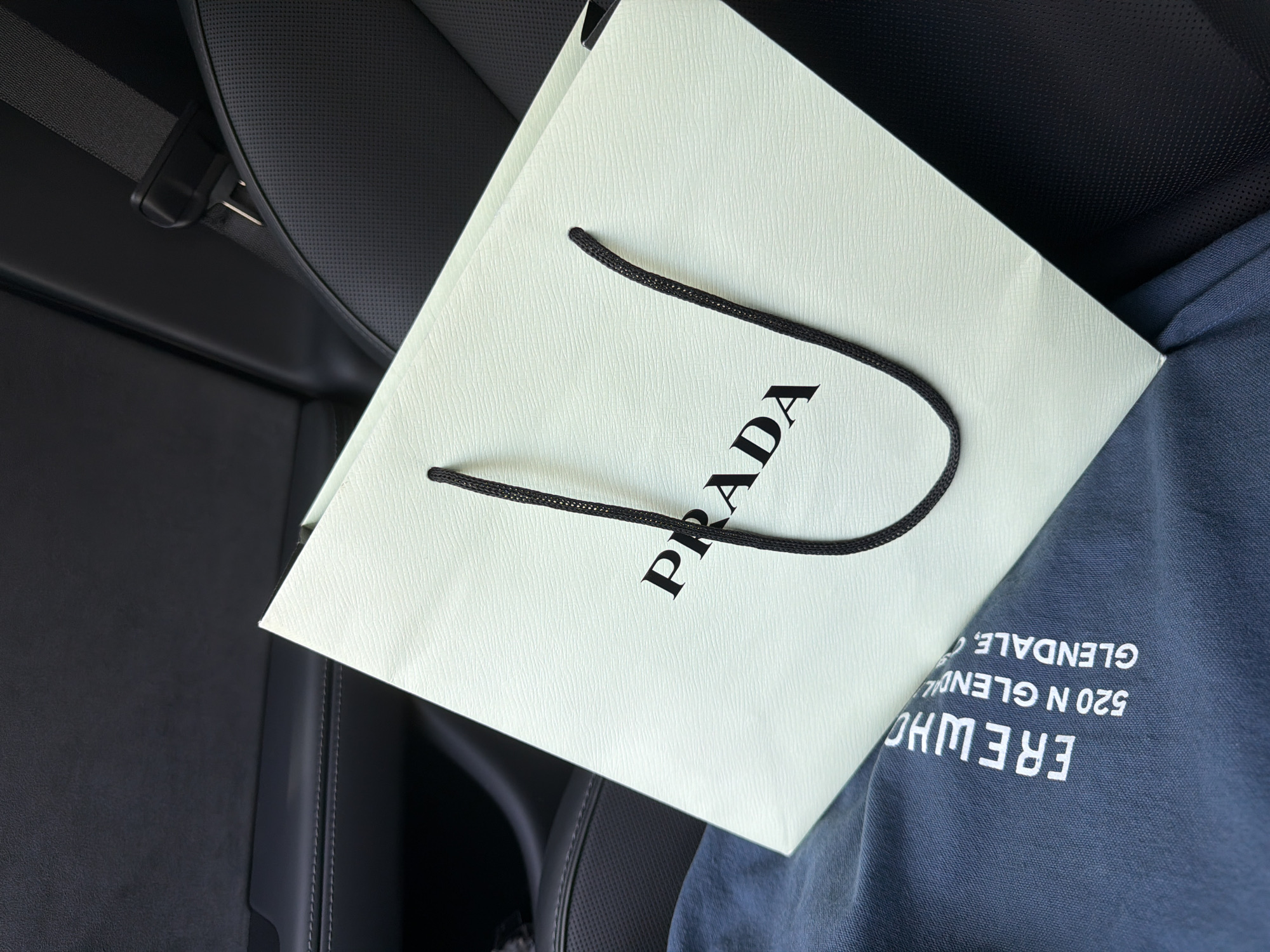 The image shows a white Prada shopping bag with black handles and the Prada logo printed on it. The bag is placed on a dark blue garment, possibly clothing, which has white text printed on it. The text reads "ERE WHO 520 N GLENDALE, C". The scene appears to be inside a car, with a black leather seat and a seatbelt visible in the background. The lighting suggests it could be daytime. The overall impression is one of luxury and shopping.