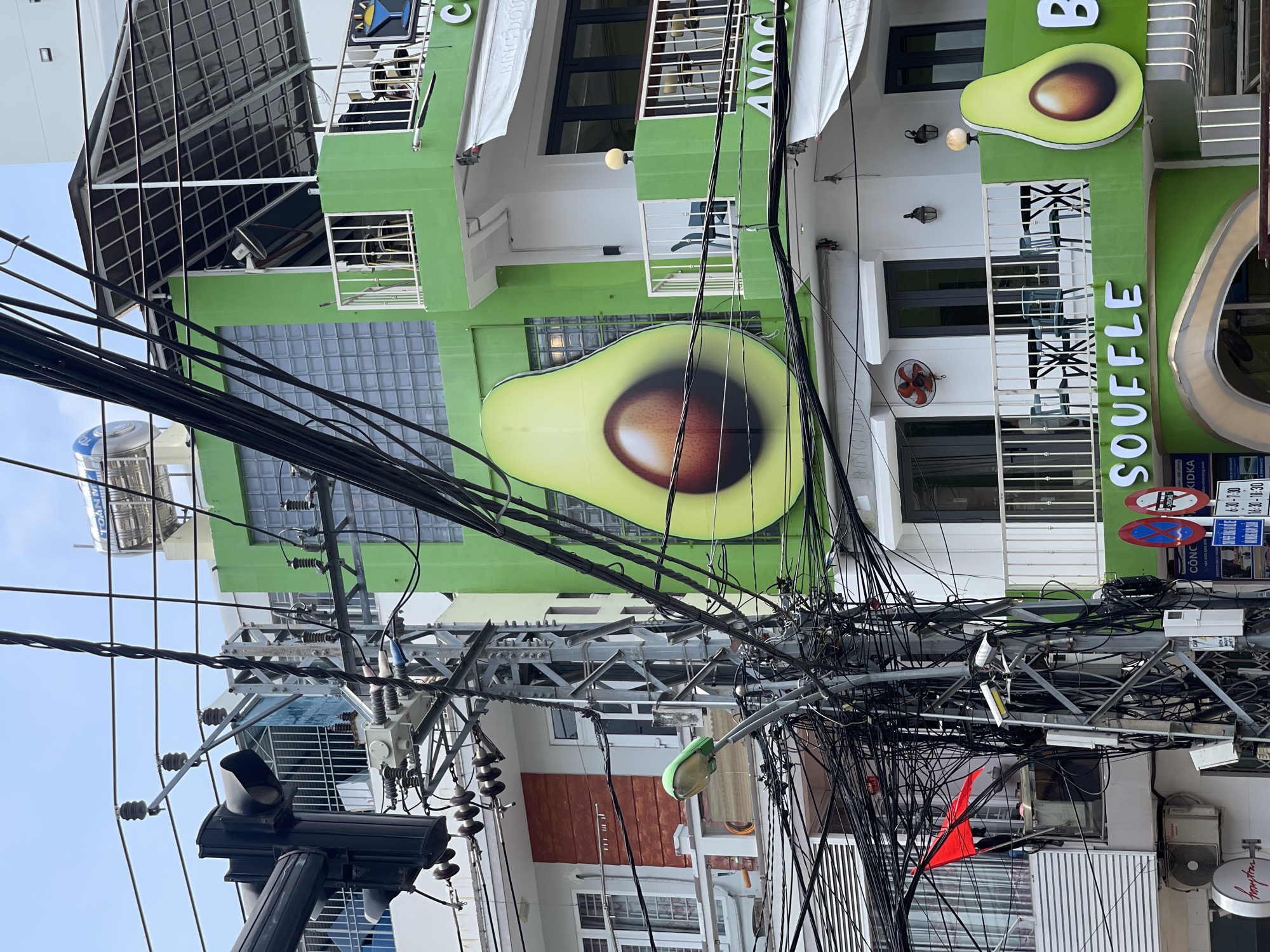 The scene captures a street view in Nha Trang, Vietnam, notable for its dense overhead tangle of power and communication lines. A vibrant, two-story building serves as the backdrop, painted in shades of green and adorned with large avocado-themed signs and the name "AVOC SOUFFLE". The building houses businesses, as indicated by signage reading "BING DOU", "C", and "Hampton".
In the foreground, a utility pole supports a complex web of black wires. Additional street furniture includes a black traffic signal head and a partially visible red flag or marker. The lower parts of buildings and storefronts are visible beneath the power lines, hinting at the urban density of the area. The photograph is taken from a lower angle, emphasizing the height of the building and the disarray of the wires against a light blue sky.