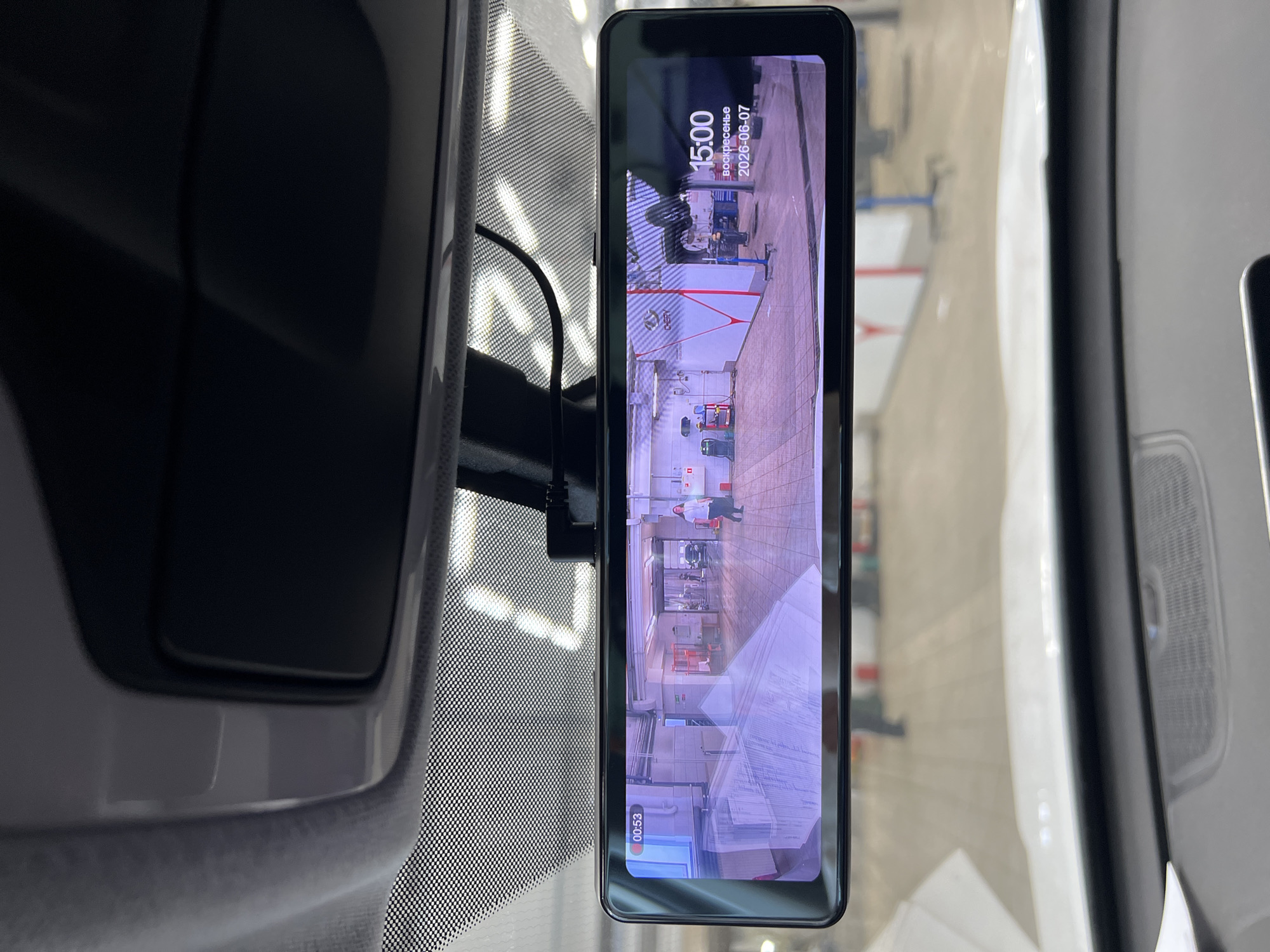 The image displays the interior perspective from a vehicle, focused on a digital rearview mirror functioning as a display for a camera feed. Above the mirror, the car's headliner and an overhead console are visible in gray and dark gray tones, respectively. The top edge of the windshield exhibits a dotted frit band.
The digital rearview mirror screen occupies the central portion of the image. It presents a wide-angle view of a car service center, identified as a Chery facility by the "CHERY" logo prominently displayed on a red and white striped wall. The service center, located in Izhevsk, Russia, features light-colored tiled flooring, pale walls, and ambient overhead lighting. Visible equipment includes a red mobile tool cart near the left wall and blue automotive lifting apparatus or stands towards the right. One adult female, dressed in a light-colored top and dark pants, is observed walking across the service bay from left to right. Overlaid on the display are a timestamp "00:53" in the upper left corner, and in the upper right, the time "15:00," the day "воскресенье" (Sunday), and the date "2026-06-07." Reflections of interior vehicle elements, possibly documents, are visible on the display's surface.