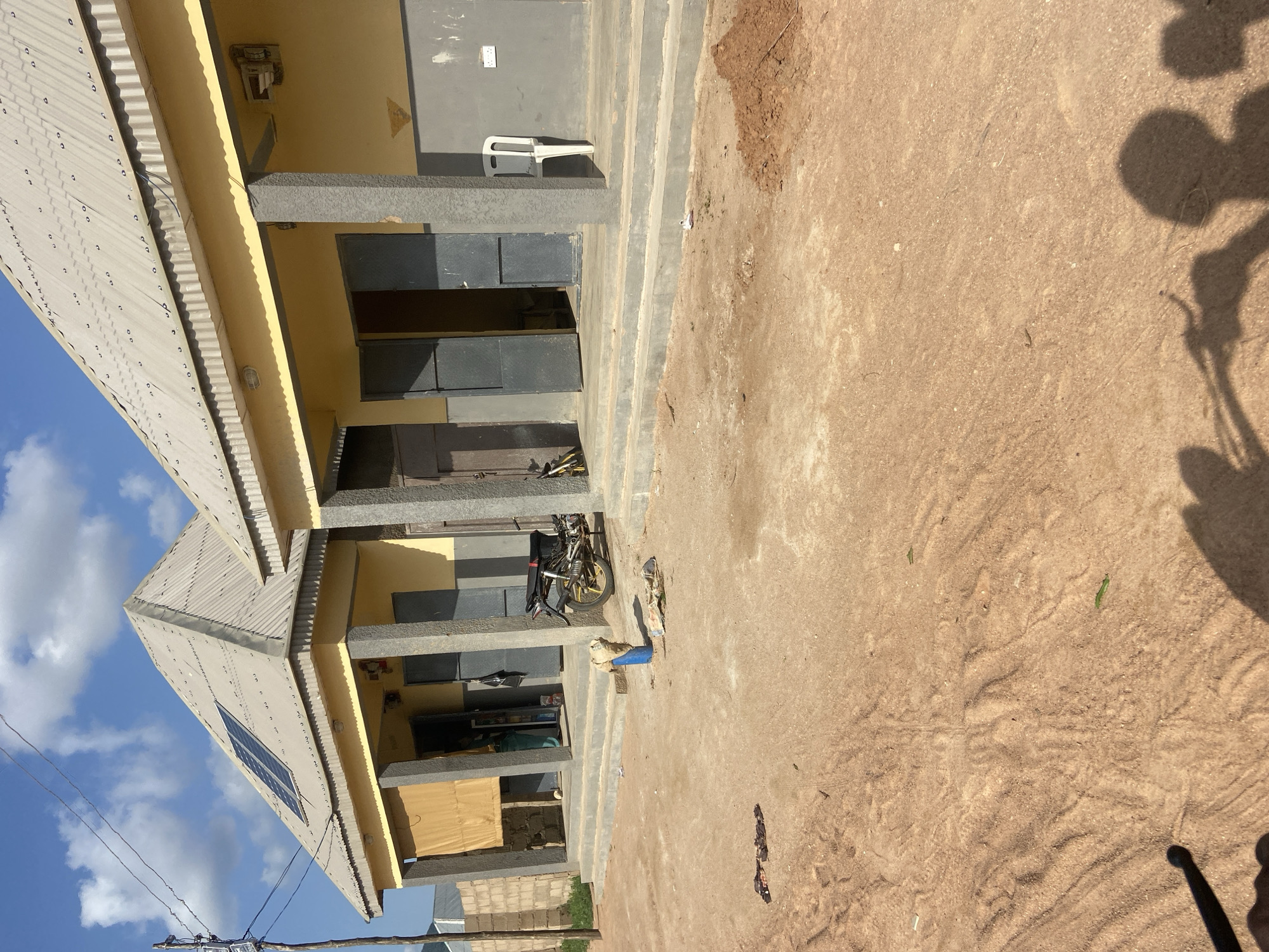 This media file shows an exterior view of a yellow building with a corrugated metal roof and a solar panel. The building appears to be a single-story structure with several entrances. In front of the building, there is a sandy, unpaved area with some scattered debris. A motorcycle is parked in front of one of the entrances. The scene suggests a warm, sunny day, with a few clouds in the blue sky. The overall impression is one of a simple, functional building in a somewhat rural or developing area, possibly a school or community center in Kuson, Nigeria. There is no discernible activity or event taking place; the image seems to be a still shot of the location.