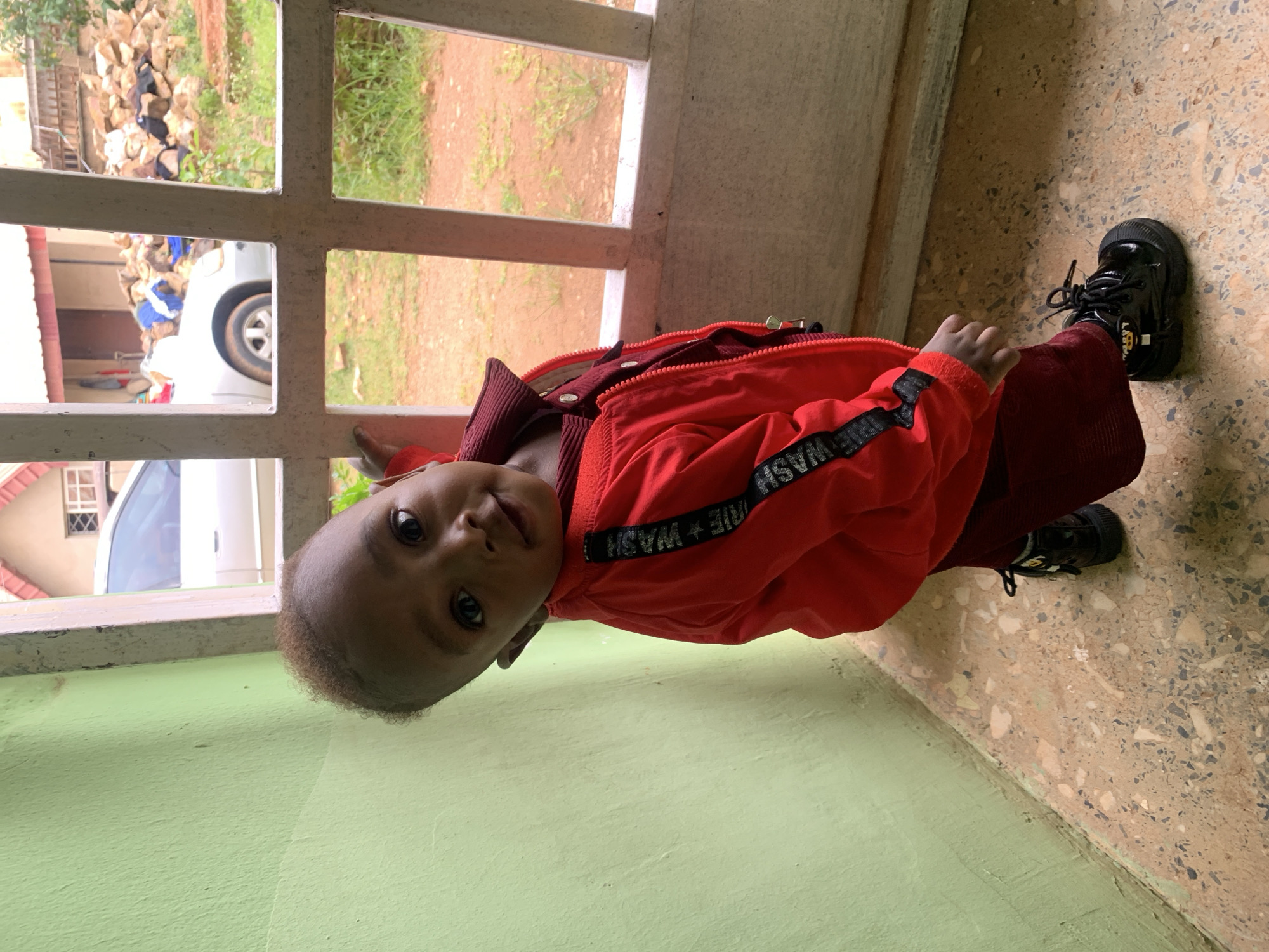 The image captures a young child standing in what appears to be a porch or entryway. The child is facing the camera and looking slightly to the side, with a curious expression. They are wearing a vibrant red jacket over a maroon outfit and black shoes. The jacket has black trim with white lettering on the sleeves.
The background suggests an outdoor or semi-outdoor setting. To the left, there is a light green wall. To the right, a white-painted metal grate or window frame divides the view of the exterior. Through the grate, a silver car is partially visible, along with a pile of rocks and some vegetation, indicating a rural or semi-urban environment. The ground in the foreground is a tiled surface with a speckled pattern.
Without further context, pinpointing the exact location in Barakin Akawo, Nigeria, is not possible from the image alone. However, the scene depicts a common residential setting. The presence of a car, building structures, and natural elements suggests a community environment.