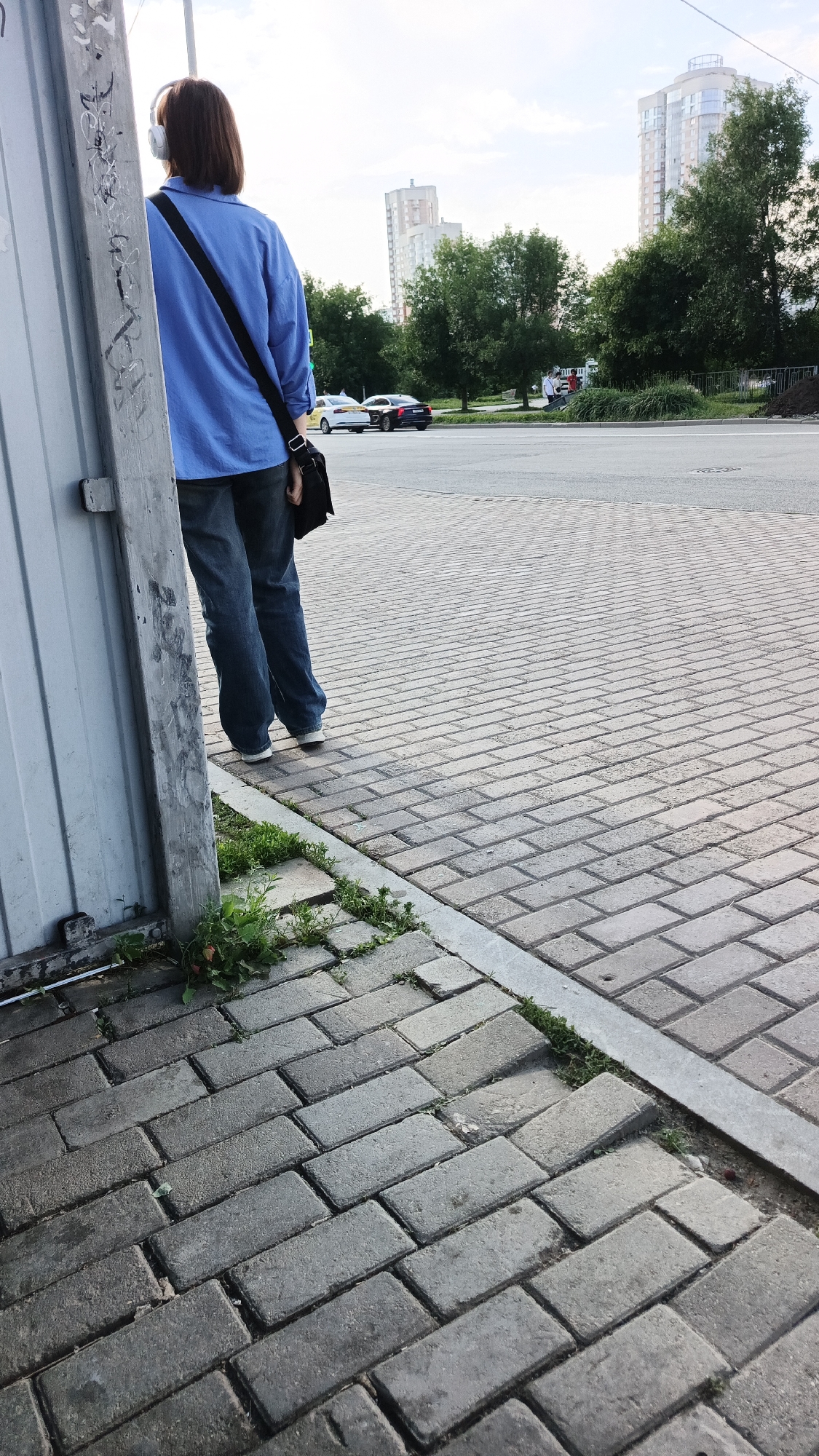 A person with short, brown hair, wearing white headphones and a bright blue collared shirt over blue jeans, stands with their back to the camera. They are leaning against a corrugated metal wall, with a black messenger bag slung over their right shoulder. The person is standing on a paved walkway made of rectangular grey bricks. A thin strip of concrete or metal separates the walkway from the street.
In the background, cars are visible on a road. One car is white, and another is dark, possibly black. Beyond the road, there are green trees and modern, tall buildings. The sky is partly cloudy, suggesting it is daytime, likely late afternoon or early evening. The overall scene appears to be an urban environment, possibly a bus stop or a street corner in Yekaterinburg, Russia. There is no visible text in the image. The person's posture suggests they might be waiting or listening to music.