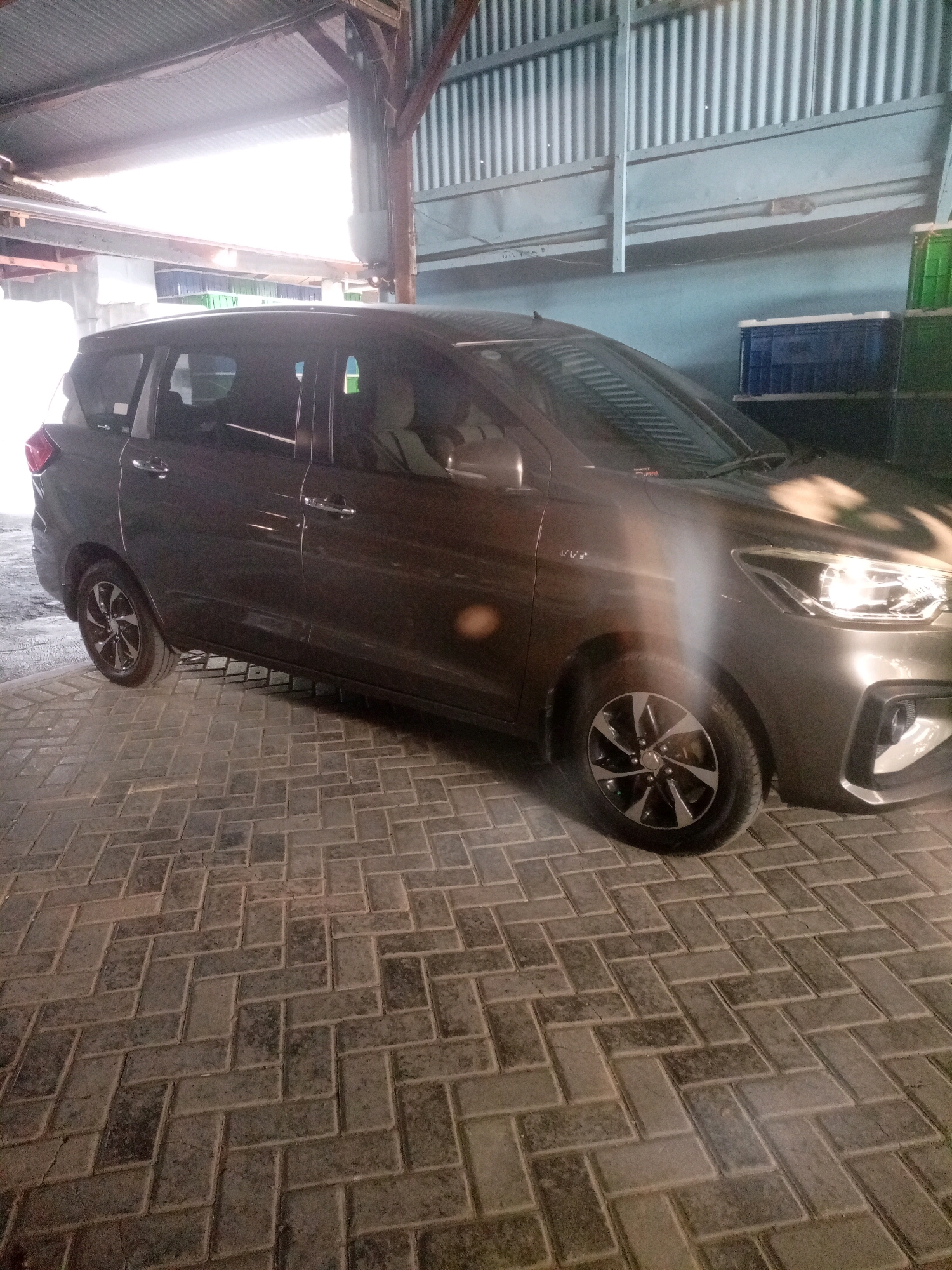 A dark gray minivan is parked on a herringbone-patterned brick driveway under a corrugated metal roof. The vehicle is facing right, with its headlights on. The scene appears to be an outdoor covered area, possibly a carport or a workshop, with a corrugated metal wall in the background. There are some green and blue crates stacked in the background, to the right of the vehicle. The floor is made of interlocking rectangular bricks laid in a staggered pattern. The lighting suggests it might be dusk or an overcast day, with some light filtering in from the left side of the image.