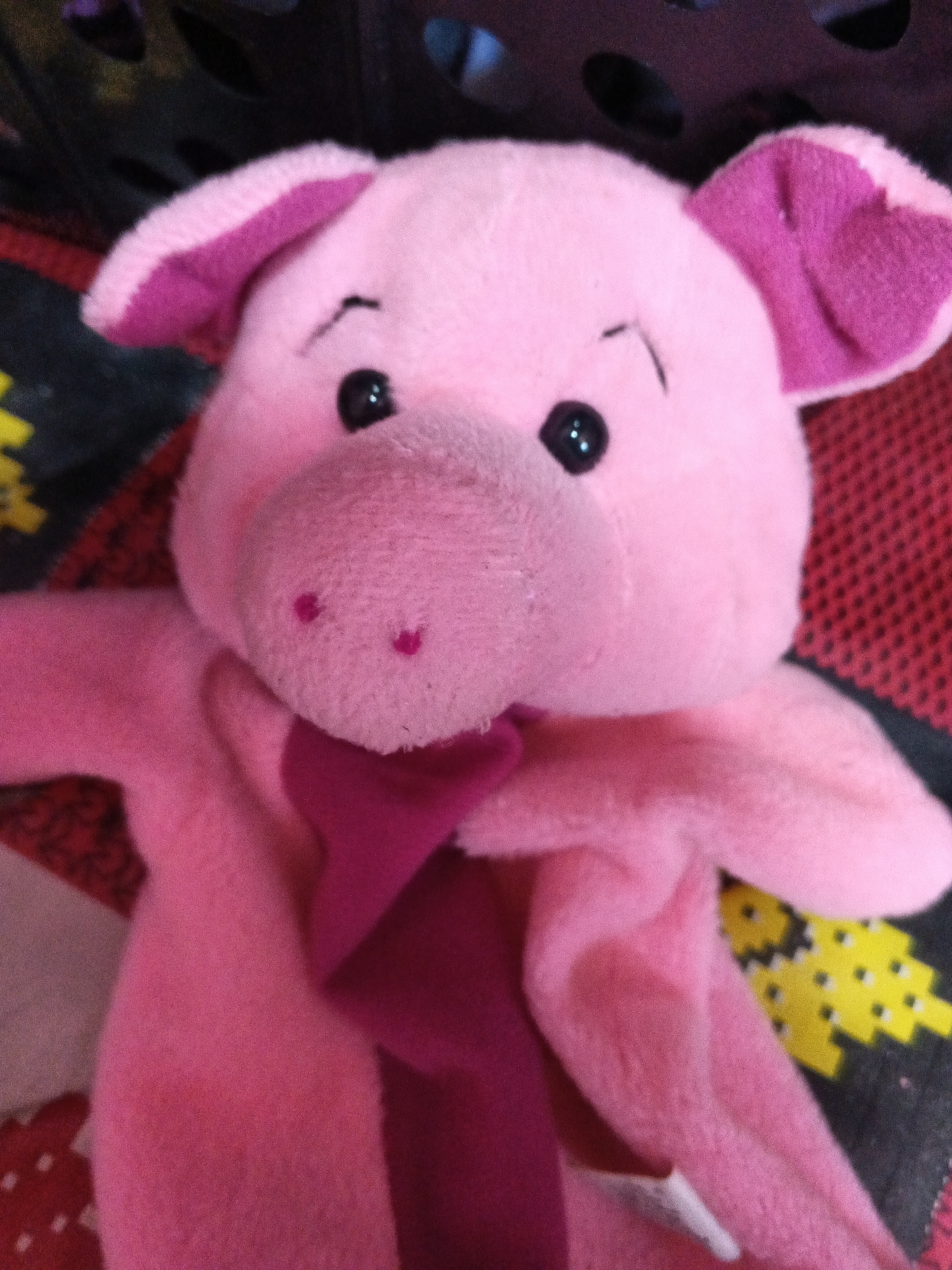 A close-up, eye-level, indoor shot shows a pink plush pig toy. The pig is held upright and facing forward. It has large, floppy ears with a darker pink interior, two black button eyes, and a small, stitched black mouth. Its snout is rounded and pink, with two darker pink nostrils. A magenta scarf or bow is draped around its neck. The pig is lying on a textured surface with a black, red, and yellow pixelated pattern. The background is dark and out of focus, with hints of a black basket weave. The lighting is dim, with some reflections visible on the pig's eyes. The scene is static and there are no people present.