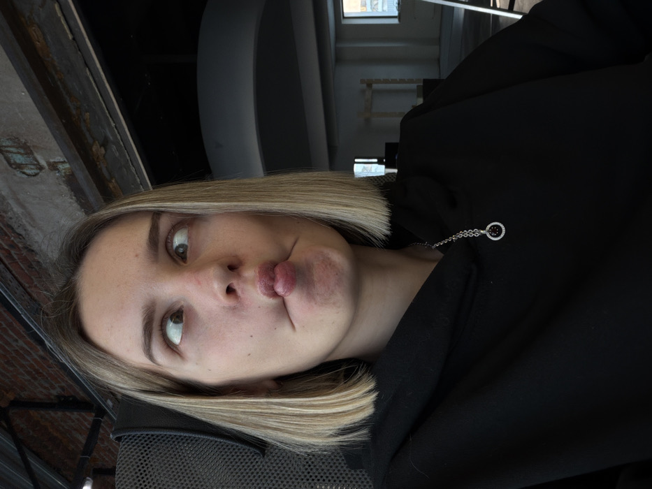 A woman with blonde, shoulder-length hair pulls a pouting face, her lips pursed in the center. She wears a black hooded sweatshirt adorned with a silver necklace featuring a round pendant. The background includes a window frame and ceiling architecture, suggesting an indoor setting. The image is shot from a slightly high angle with a rotated perspective.