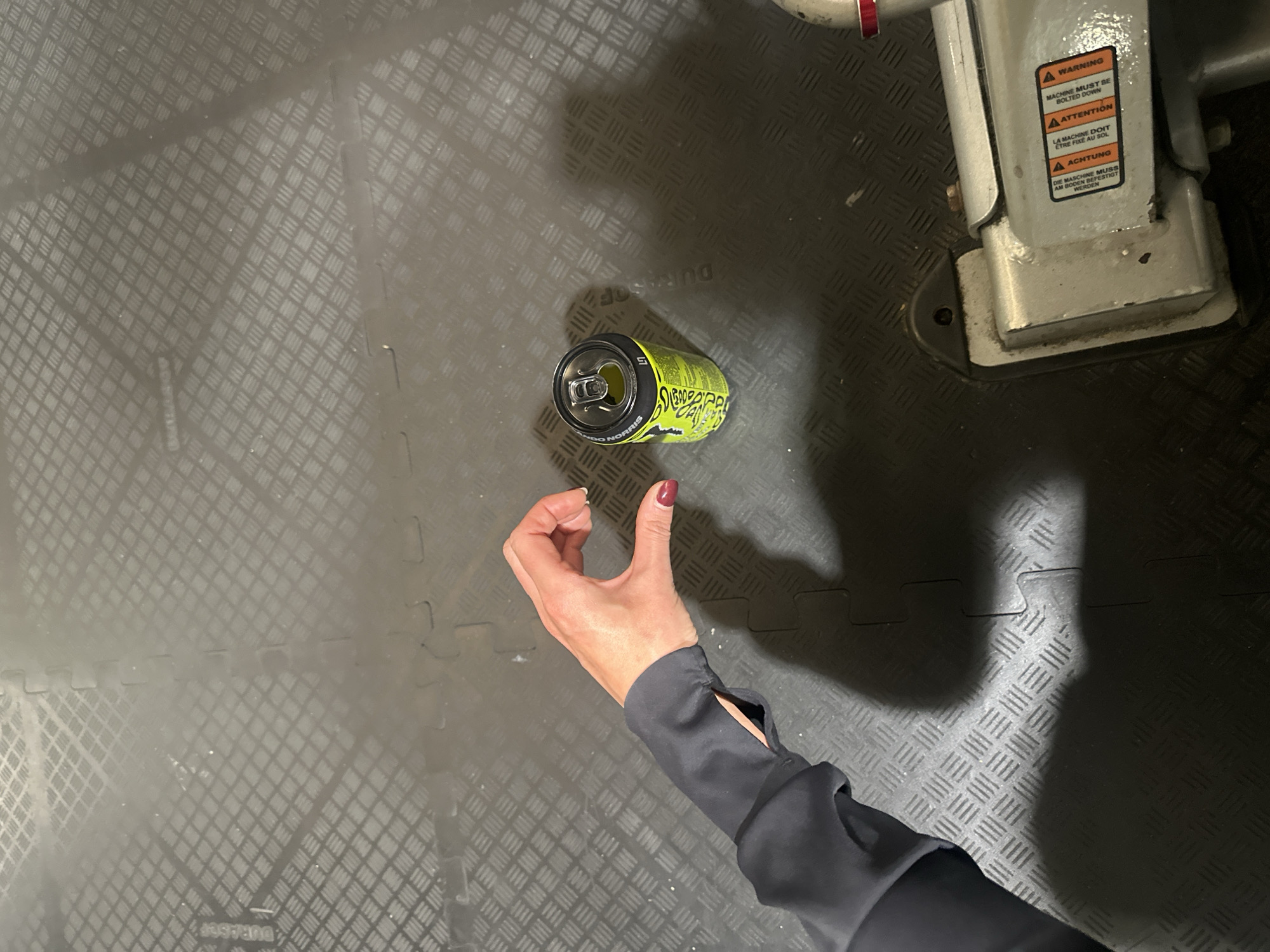 The image shows an overhead shot of a floor, likely in an industrial or workshop setting, given the checkered pattern and the presence of what appears to be a piece of machinery in the upper right corner, labeled with warning signs in English, French, and German. A hand is visible, partially obscuring a green and black energy drink can labeled "Chuck Norris." The floor appears to be made of rubber or a similar material, likely for durability and slip-resistance. There is no indication of the location being in Tunbridge Wells, United Kingdom.