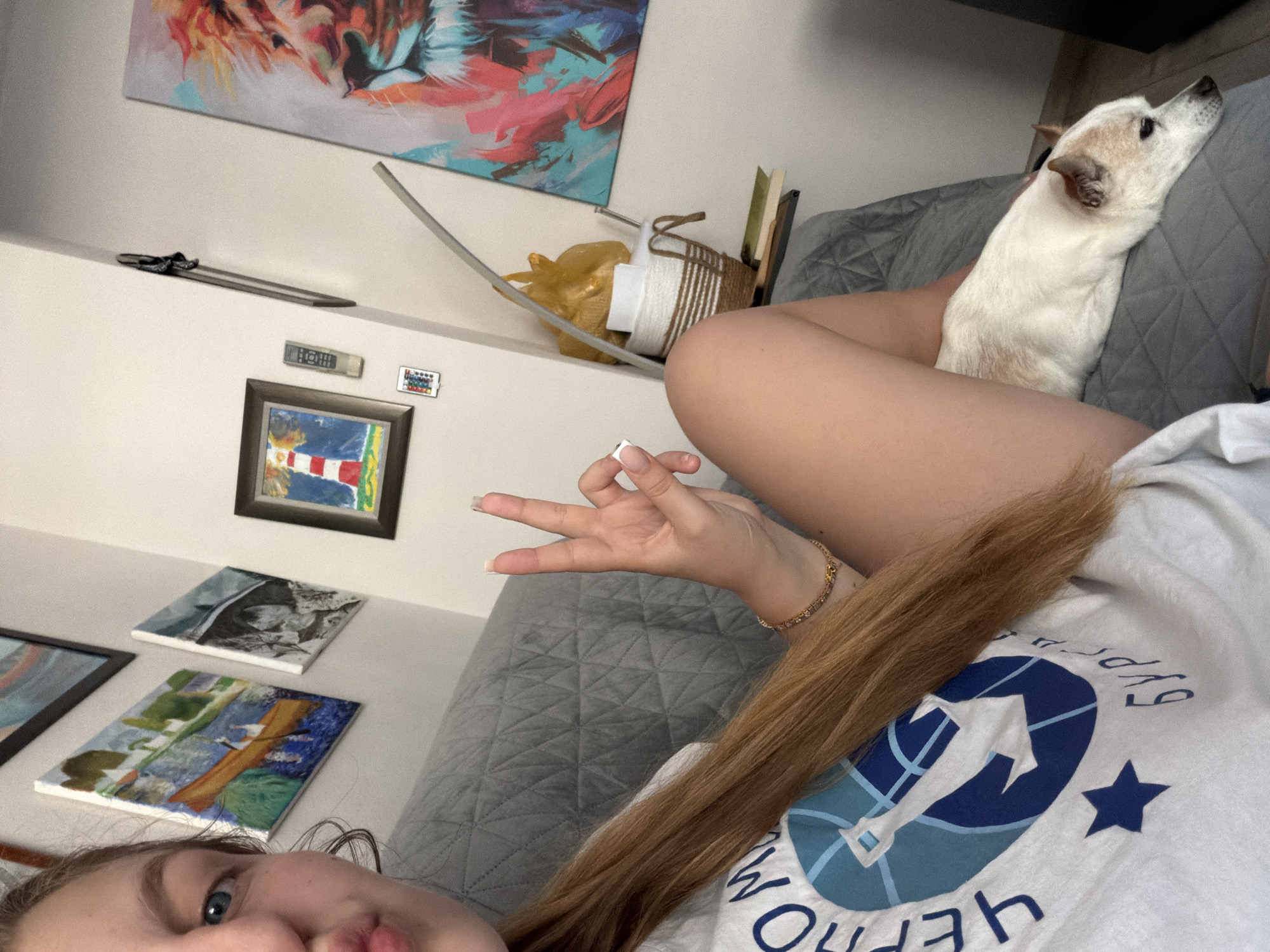 The image captures a close-up of a young woman with long blonde hair, resting on a grey sofa in what appears to be an indoor setting, possibly an apartment. She is wearing a white t-shirt with a blue logo that includes text in Cyrillic script, which reads "ЧЕРНОМОРЕЦ БУРГАС," indicating a connection to the Chernomorets Burgas sports club, suggesting the location is indeed Burgas, Bulgaria. The woman is making a peace sign with her right hand. Beside her, a small white and tan dog is lying down on the sofa. In the background, the wall is adorned with several framed paintings, including one of a lighthouse and another of a lion's head. A small shelf holds a woven basket, some books, and a yellow plastic bag. A remote control and a smaller decorative item are also visible on the wall. The overall scene conveys a relaxed, casual atmosphere.