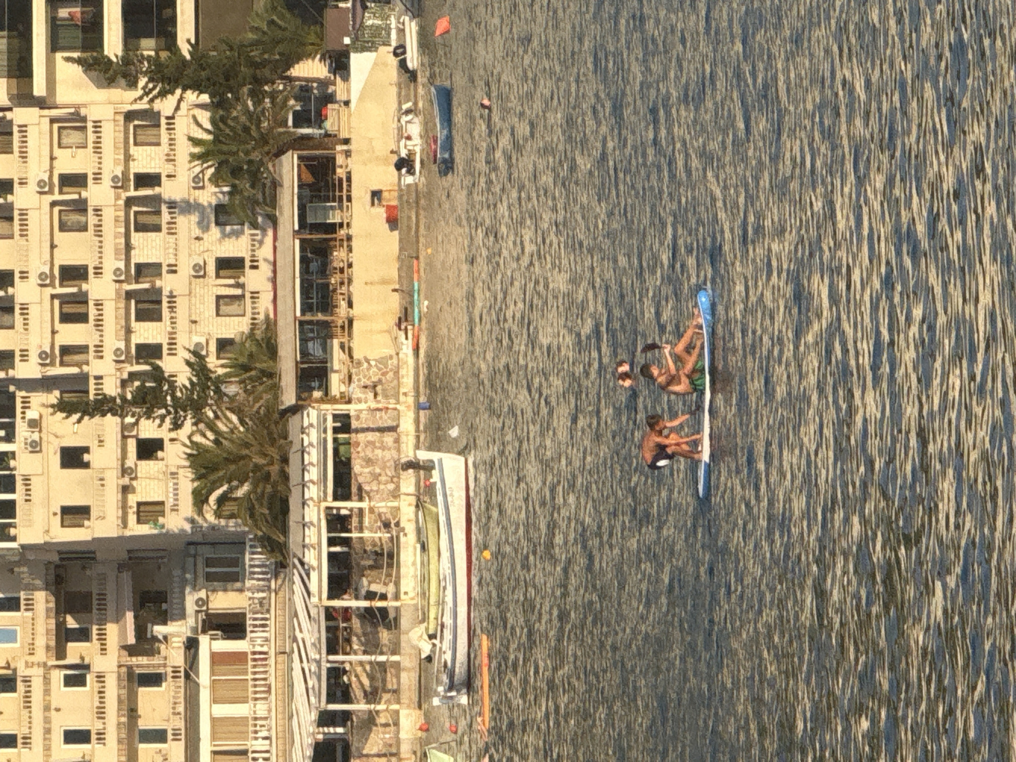 In the coastal city of Rafailovići, Montenegro, a tranquil scene unfolds on the water. The foreground is dominated by a stand-up paddleboard with three individuals. One person, wearing dark shorts, kneels on the board, actively paddling with a dark oar. Beside them, another individual, wearing green shorts, sits upright, while a third person leans back on the board. The group appears to be enjoying a leisurely outing on the water.
In the middle ground, several boats are anchored or moored. A long white boat with a red stripe is prominently positioned, with another lighter-colored boat nestled behind it. Further in the distance, a small blue boat is visible. The water itself is calm, reflecting the golden light of what appears to be late afternoon or early morning.
The background features a multi-story building with numerous balconies and windows, characteristic of a coastal resort. Adjacent to the building is a restaurant or beach bar with an outdoor seating area, partially covered by a white awning. Palm trees and other greenery are interspersed around the structures, adding to the Mediterranean ambiance. The scene captures a moment of relaxation and recreation by the sea, with the architecture and natural elements creating a picturesque backdrop.