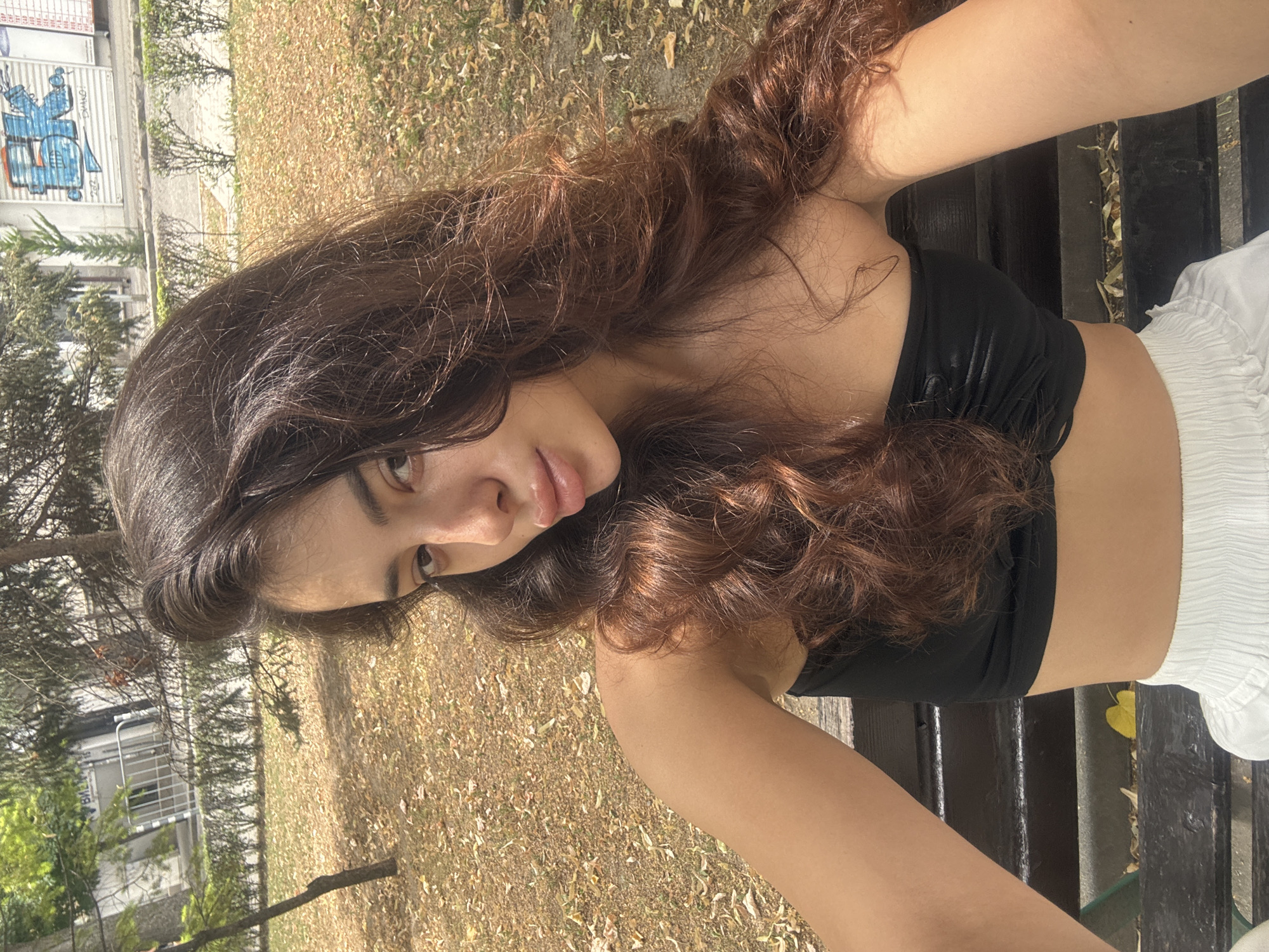 The media file shows a young woman with long, wavy, dark brown hair, sitting outdoors on a dark wooden bench. She is wearing a black tube top and white ruffled shorts. The woman is looking towards the camera with a soft expression. Her face is partially obscured by her hair. Sunlight illuminates her skin and hair.
The background consists of dry, fallen leaves on the ground, suggesting an autumn setting. To the left of the woman, there are trees with bare branches and some greenery. In the far left background, a white building with a metal fence and graffiti can be seen. The graffiti appears to be a blue stylized robot or figure. There are also some horizontal red and white signs on the building. The lighting suggests it is daytime, likely late morning or afternoon, given the warm, direct sunlight. The overall mood is relaxed and casual.
The location context provided is Plovdiv, Bulgaria, which aligns with the outdoor setting and architectural style of the building in the background. No specific event or activity is clearly depicted, other than the woman posing for a photograph.