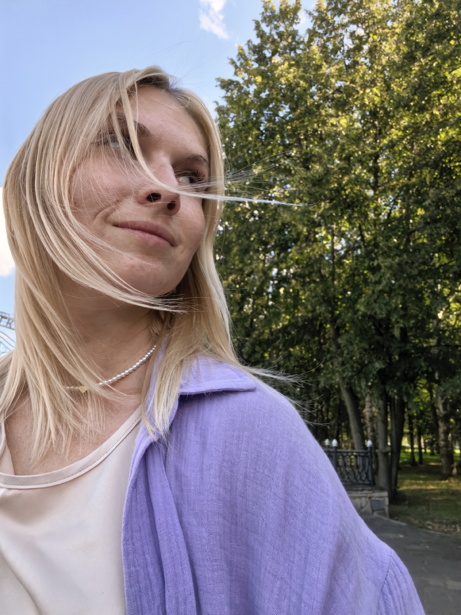 A young woman with blonde hair and a pearl necklace smiles as she looks to the right. She is wearing a light-colored tank top and a lavender shirt. Her hair is blowing in the wind. Behind her is a park with green trees and a blue sky with some clouds.