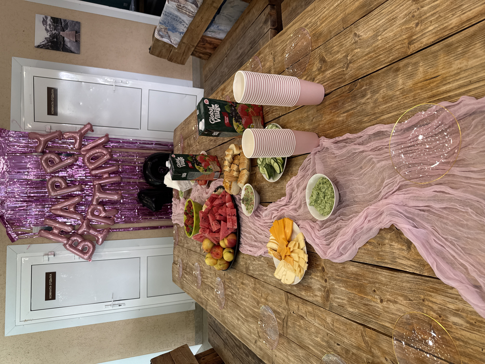 This media file shows a wooden table set up for a party, likely a birthday celebration given the "HAPPY BIRTHDAY" balloons visible. The table is laden with food and drinks, including a platter of sliced watermelon and apples, a cheese board with various cheeses, bowls of green dip or salad, and sliced bread. There are also two cartons of Global Village juice, one featuring berries and another with mixed fruits. Pink paper cups are stacked on the table, along with clear plastic plates with gold rims. A pink, gauzy fabric drapes across the table, adding a festive touch. In the background, two white doors with text are visible, one of which reads "КОМНАТА ОТДЫХА" (Rest Room). The overall impression is one of a casual, indoor gathering. The lighting suggests it could be daytime, and the setting appears to be a room with a somewhat rustic decor, possibly a cafe or a recreational area in Chaikovsky, Russia.