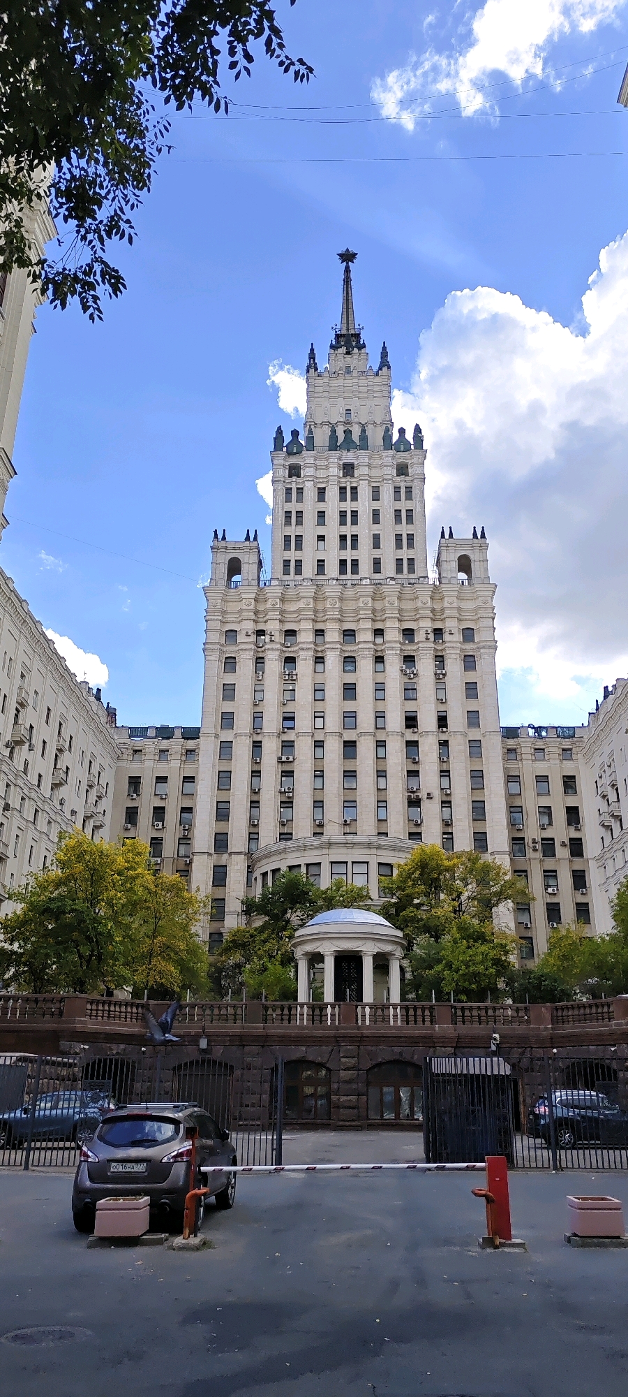 This image depicts a tall, ornate apartment building in Moscow, Russia, with a distinct spire topped by a star. The building is constructed of light-colored stone and features numerous windows arranged in a grid pattern. Several trees with green and yellow leaves are visible in front of the building, and a small, white gazebo-like structure with a blue roof is situated in the middle ground. The foreground shows a street with a dark asphalt surface, a barrier arm, a red bollard, and two parked cars. A pigeon is in flight near the barrier. The sky is blue with scattered white clouds.
