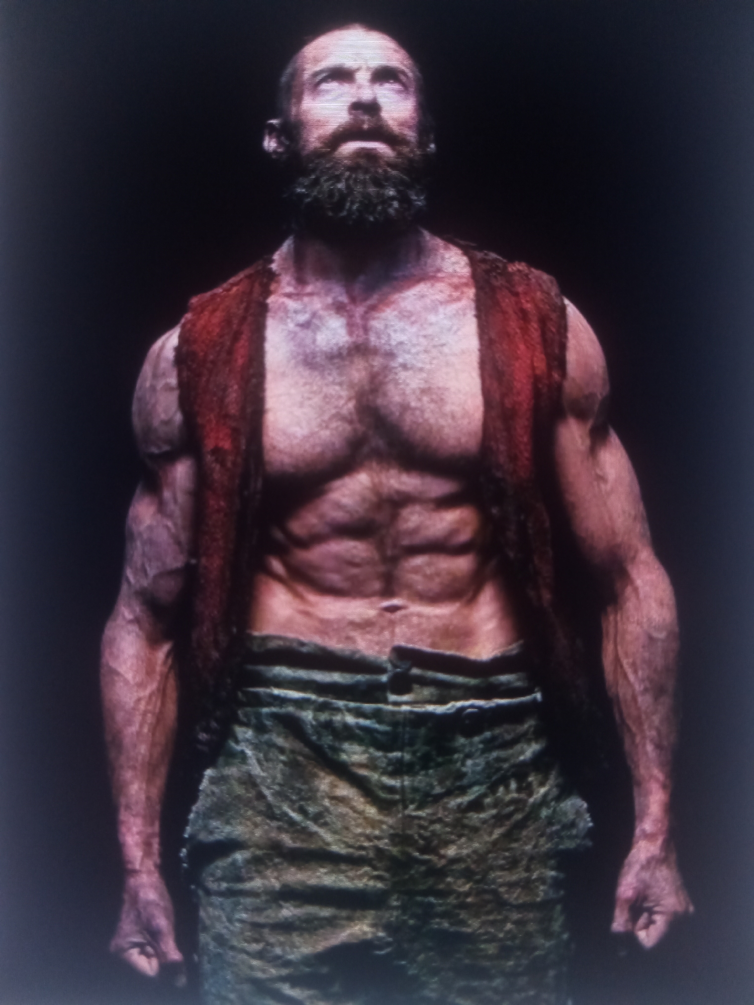 A low-angle, full-body shot shows a muscular man with a beard looking up against a dark background. He wears a red vest over his bare chest and a rough, textured garment around his waist. His skin appears reddish and bruised, particularly on his chest and arms, with prominent veins visible. The man's physique is highly defined, showcasing his abdominal and arm muscles.