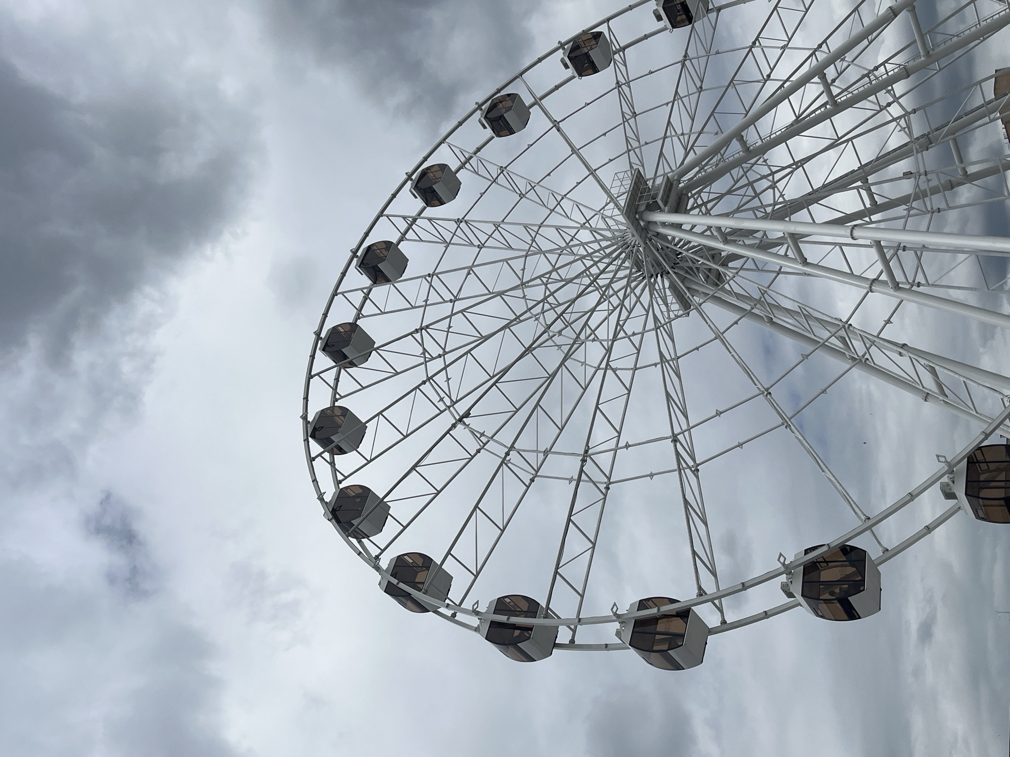 The image shows a Ferris wheel against a cloudy sky. The Ferris wheel is white and has many gondolas. The gondolas are enclosed and appear to have tinted windows. The structure of the Ferris wheel is intricate and made of metal beams. The sky is filled with dark, gray clouds, suggesting overcast weather.
The Ferris wheel is located in Saratov, Russia. It is a prominent landmark in the city and a popular attraction for both locals and tourists. The specific location in Saratov is on the embankment of the Volga River, offering panoramic views of the city and the surrounding landscape. The Ferris wheel is part of the "Victory Park" (Park Pobedy) amusement area.
