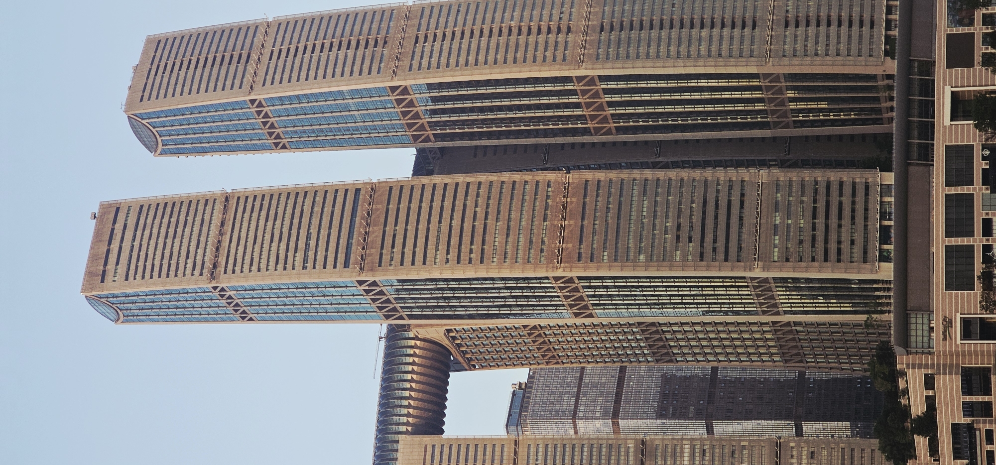 The image shows a close-up, low-angle view of several modern skyscrapers in the Nan'an District of Chongqing, China. The skyscrapers are characterized by their distinctive architecture, featuring a combination of glass facades and horizontal beige panels, creating a ribbed or layered appearance. The two most prominent towers are tall and slender, with one on the right slightly wider than the one on the left. Diagonal cross-bracing is visible on the exterior of these towers. To the left, a U-shaped structure connects two buildings, resembling a large, open archway or a bridge. Below this archway, another building with a darker, reflective facade is visible. At the very bottom of the frame, a lower, multi-story building with large rectangular windows and beige horizontal banding is partially visible, suggesting a commercial or retail area. The sky is clear and a pale blue, indicating daylight. No people or specific activities are discernible within the scene.