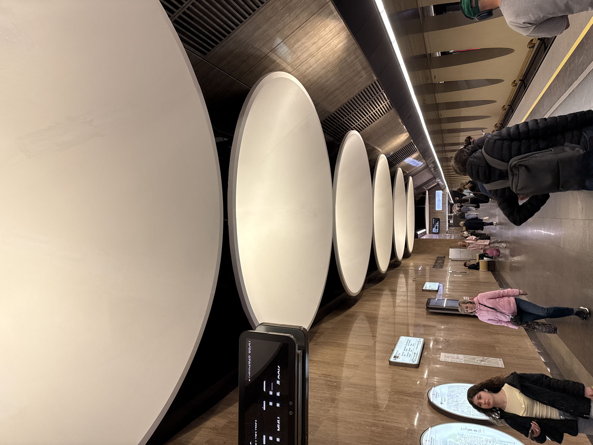 The image shows a view within the Park Pobedy Metro Station in Moscow, Russia. A series of large, white, oval-shaped art installations line one side of the station platform. The platform itself is made of polished stone. Digital information displays and map kiosks are scattered along the platform floor. People are present on the platform: some are standing, some are walking, and some are lying down on benches. The ceiling above is a combination of textured panels and linear lighting fixtures. A person is holding a smartphone with the screen facing the camera.
