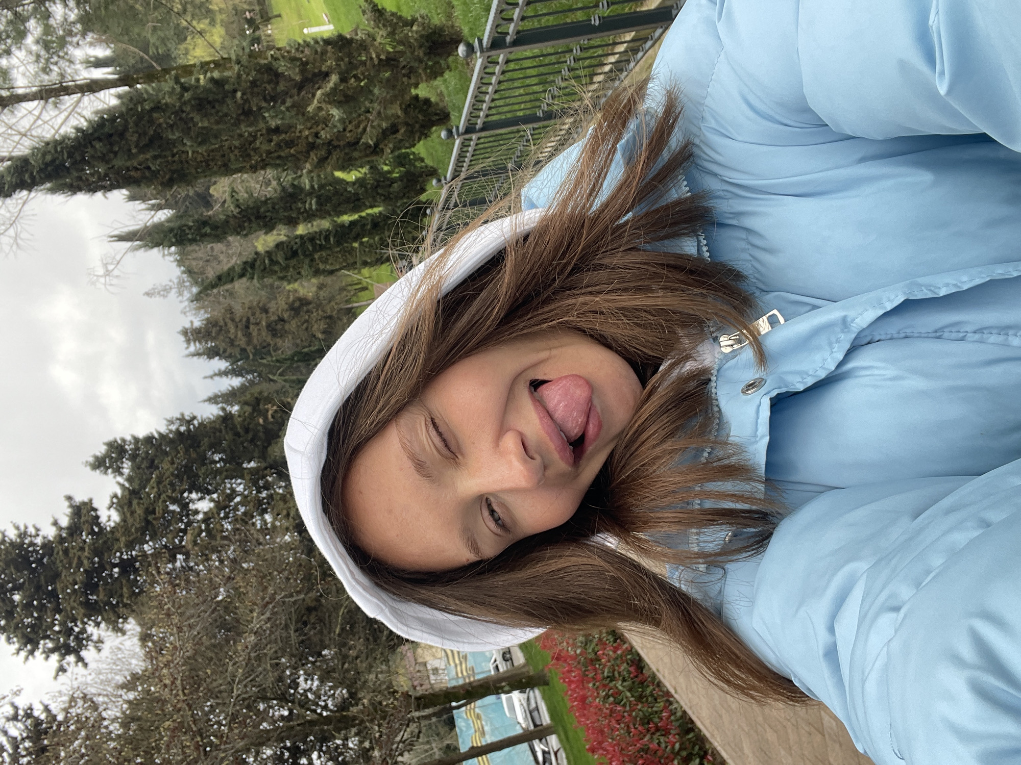 A young female is depicted in a close-up selfie, occupying the majority of the frame. She has light brown hair, partially covered by a white hood. She is wearing a light blue puffer jacket with a visible zipper and snap button closure. Her facial expression includes her right eye closed in a wink and her tongue extended from her mouth. Her skin shows a scattering of light freckles.
The background is an outdoor environment in the city of Sochi, Russia. To the left, a paved pathway runs alongside a low hedge of red-leafed plants. Beyond this, a road is visible with at least two parked vehicles, including a white SUV, and a blue and yellow patterned barrier or structure. Deciduous trees with bare branches stand further back. To the right of the subject, a black metal fence with decorative finials separates the path from a grassy slope. Numerous tall, slender coniferous trees, such as cypresses, populate the ascending landscape. Denser evergreen trees are visible in the distant background, under an overcast, light grey sky.