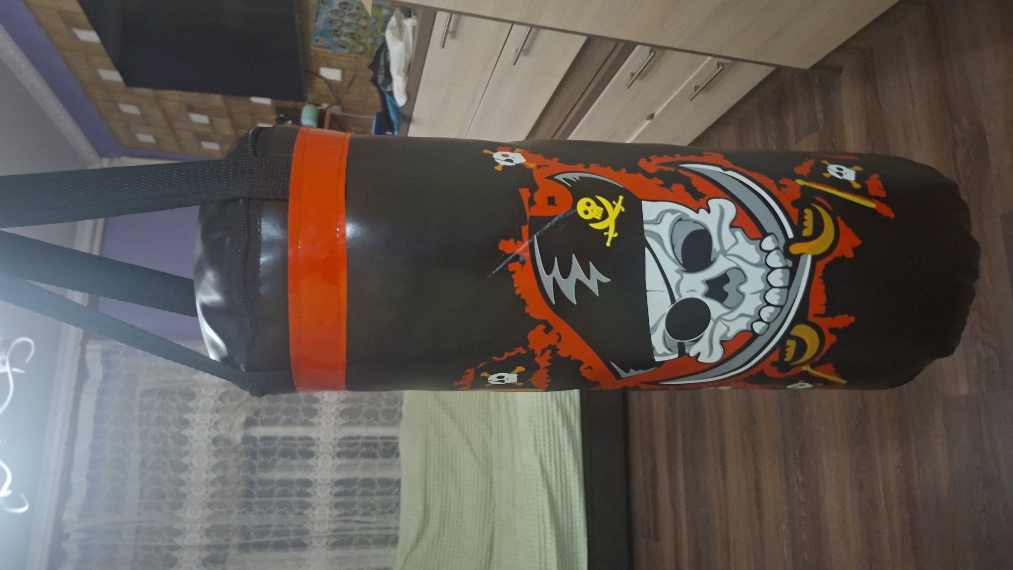 A black punching bag with a pirate skull and crossbones design is hanging in a room. The punching bag has red stripes at the top. In the background, there is a light-colored couch with a green blanket, sheer white curtains, a wooden floor, and a dresser with light-colored drawers. The overall scene suggests an indoor domestic setting.
