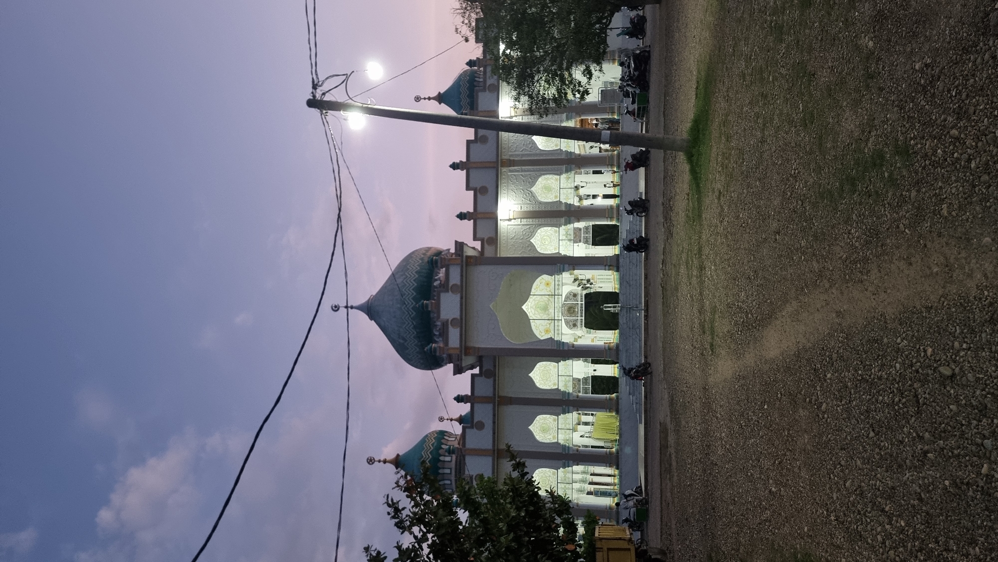 This is a low-angle, eye-level, full shot of a mosque in Lapang Timu, Indonesia, at dusk. The mosque is the central subject, featuring a large green dome and multiple smaller domes, along with ornate white arches and columns. The building is illuminated, casting a warm glow. In front of the mosque, there is a paved area with several parked motorcycles and a few people. To the left, a portion of a truck is visible, and a tree with dark green foliage partially obstructs the view of the mosque. In the foreground, the ground is covered in gravel with a dirt path winding through it. Overhead, power lines and lights are strung across the sky, which is a gradient of purple and pink, indicating the transition from day to night. The overall scene suggests a public space during a time of prayer or evening gathering.