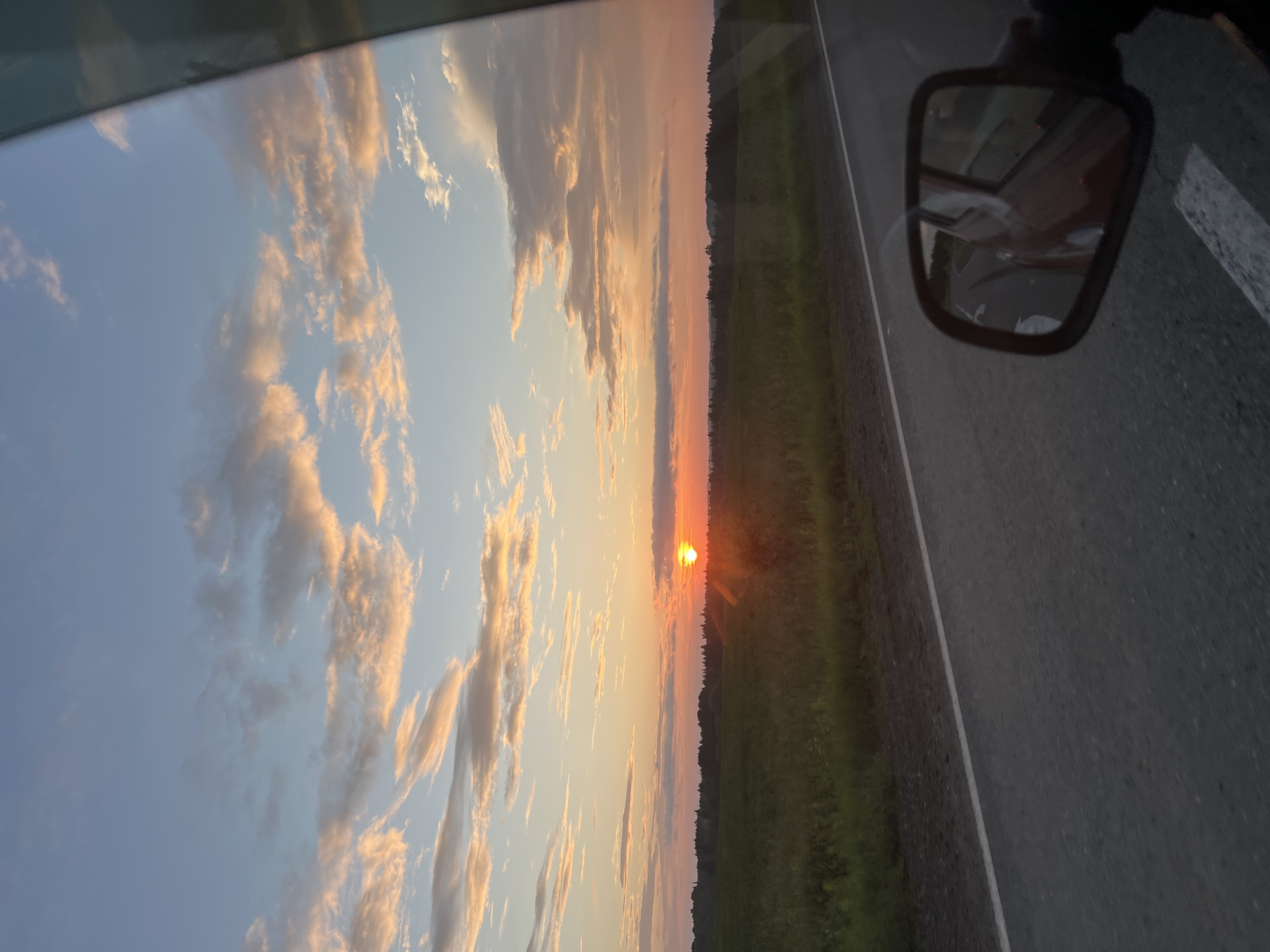 The image captures a sunset scene viewed from a moving vehicle, likely on a rural road, with a glimpse of the sky and landscape. The sky displays a gradient of colors from pale blue at the top to vibrant orange and pink near the horizon, where the sun is a bright, setting orb. Wispy, light-colored clouds are scattered across the sky, catching the warm hues of the sunset. Below the sky, a dark line of trees forms the horizon, bordering a wide, green field that extends to the foreground. A paved road with a white line marking its edge runs diagonally across the lower portion of the image. In the bottom right corner, a side-view mirror of the vehicle reflects the road behind and a portion of the vehicle's red interior. The scene does not contain any specific identifiable landmarks or elements that would place it within the city of Dokshitsy, Russia.