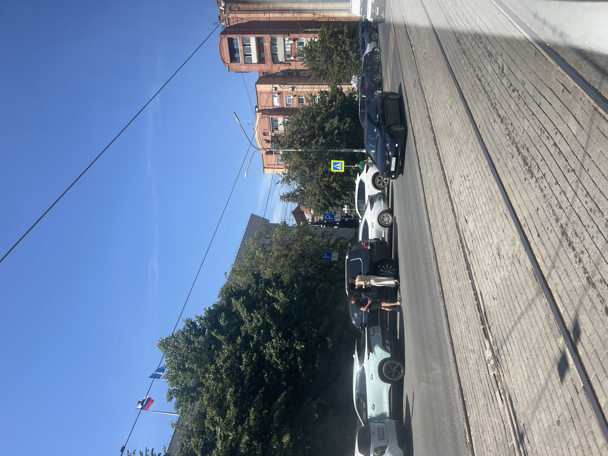 The image depicts a street scene in Ulyanovsk, Russia, likely on a sunny day, given the clear blue sky. In the foreground, tram tracks are visible on a paved walkway. A multi-lane road runs parallel to the walkway, with several cars parked or driving.
On the road, a light blue SUV is parked next to a black SUV. A man in a black t-shirt and shorts and a woman in a light-colored dress are standing between the two vehicles, appearing to interact. Other vehicles are also present, including a black sedan in motion on the road, and several parked cars of various colors and models.
In the background, a large building with trees in front of it is visible. Across the street, a red brick building with multiple floors and windows dominates the view, with a smaller brick building adjacent to it. Two flags, one red, white, and blue (likely the Russian flag) and another blue flag, are flying from a pole on top of a structure behind the trees. A yellow pedestrian crossing sign is also visible. The overall impression is of a typical urban street in Russia.