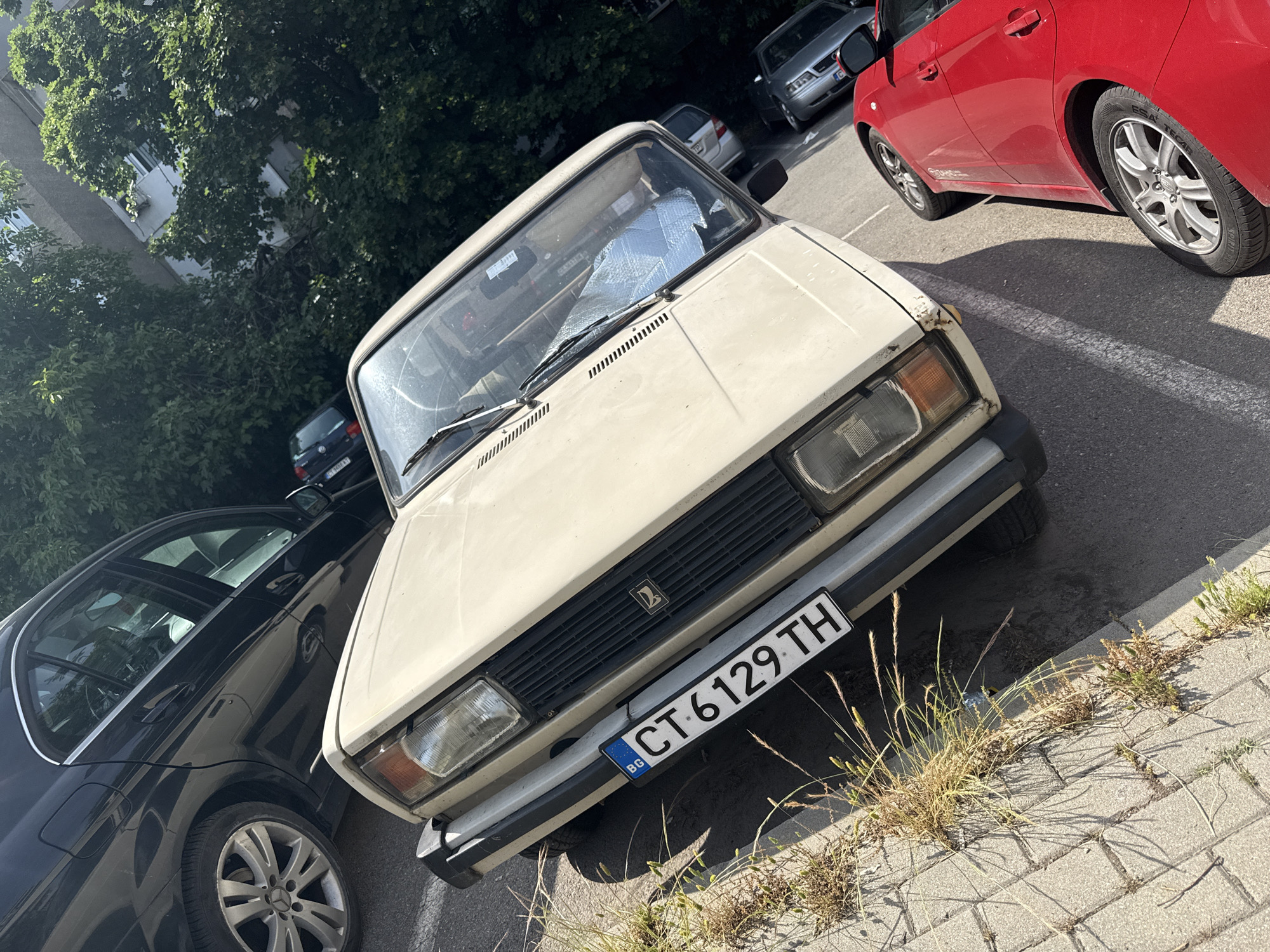 The image presents an outdoor parking scene in Stara Zagora, Bulgaria, under bright daylight. The central subject is an older, cream-colored Lada sedan, parked diagonally in the foreground. Its front features rectangular headlights, a black grille with a Lada logo, and an older, slightly worn bumper. A silver reflective sunshade is visible through its windshield.
To the right of the Lada, a portion of a red hatchback is visible, showcasing an alloy wheel with tire branding "ESA TECAR" and "SPIRIT." On the left, a black Mercedes-Benz sedan is partially seen, identifiable by its distinctive wheel design and Mercedes-Benz cap. In the background, several other cars are parked, including a silver sedan with a partially visible "CA" on its license plate, and another darker vehicle.
The setting appears to be an asphalt parking area with faint white lines, transitioning to concrete paving stones in the immediate foreground, where patches of grass and weeds grow. Lush green trees form a dense canopy in the background, partially obscuring a light-colored building with windows and an air conditioning unit.
The Lada's front license plate prominently displays "BG" on a blue strip, followed by "CT 6129 TH." The "CT" prefix on the plate confirms its registration in Stara Zagora. The clear, direct sunlight indicates daytime, likely morning or afternoon, with no specific human activity visible, only parked vehicles.