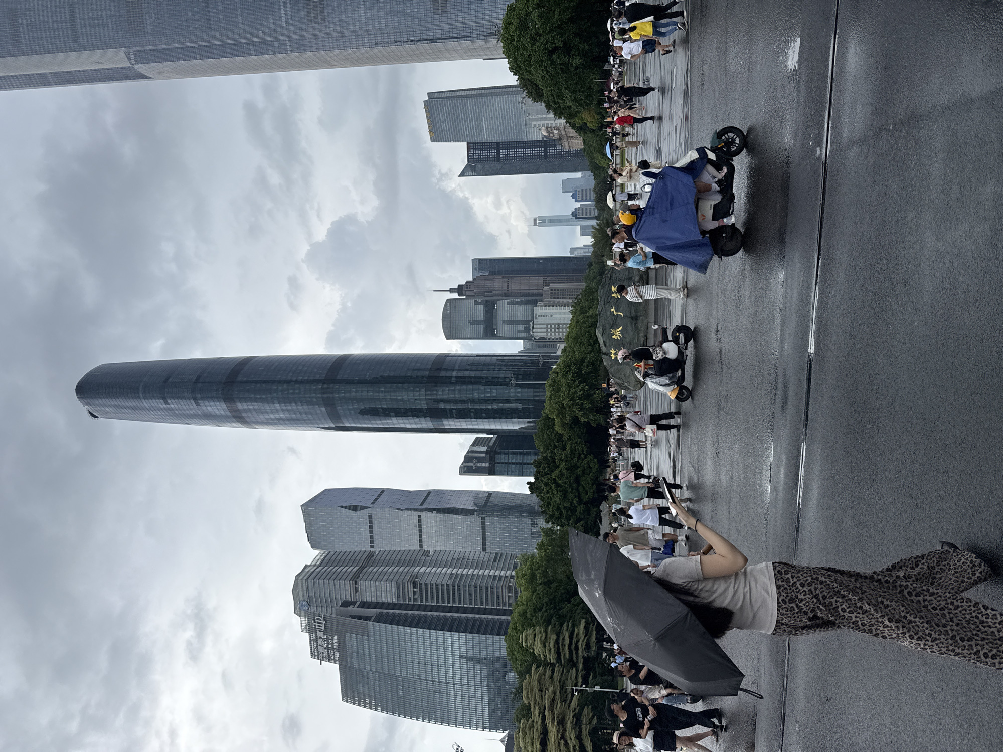 The image depicts an overcast day in Tianhe District, China, characterized by a cityscape dominated by modern skyscrapers. The foreground shows a wet asphalt road, indicating recent rain or ongoing precipitation, with several people walking and some on scooters. A woman in the immediate foreground is walking with a black umbrella and holding a smartphone. In the middle ground, a group of people are seen strolling on the wet pavement, some holding umbrellas. Two individuals are riding a scooter covered by a blue tarp. In the background, numerous tall glass-fronted buildings rise into the cloudy sky. The architecture suggests a bustling urban environment.