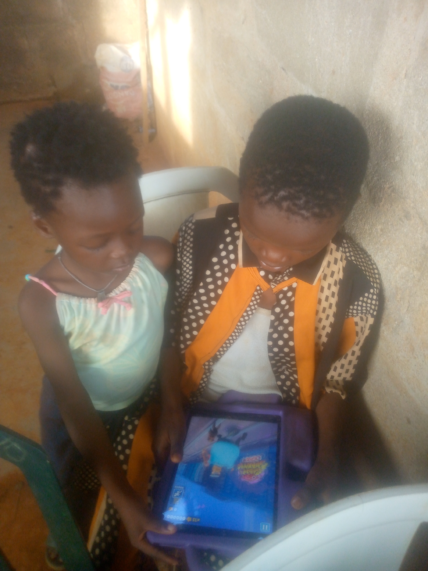 Two young children are sitting close together on a white plastic chair, likely indoors, watching a blue tablet. The girl on the left wears a light-colored tank top with pink straps and a silver necklace. The child on the right is wearing a brown and tan patterned shirt. The tablet displays a cartoon. The background includes a rough-looking wall and a bag resting on what appears to be a ledge or another chair. The location is Ikorodu, Nigeria.