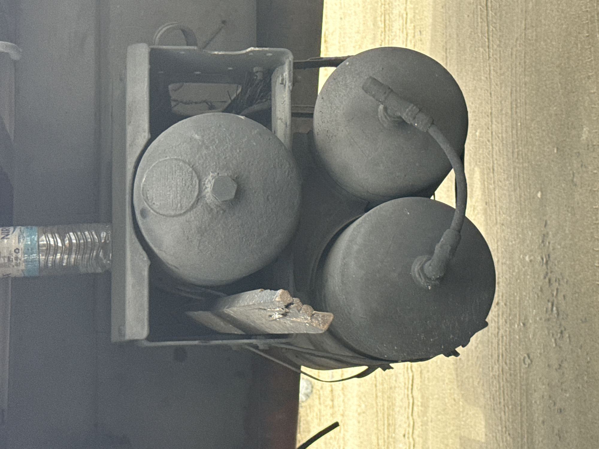 The image shows a set of three cylindrical air tanks mounted in a metal frame. The tanks are covered in dust or grime. A plastic water bottle is visible to the left, and a piece of broken wood rests beneath the tanks. The scene appears to be outdoors, with a concrete or asphalt surface visible in the background. The image is taken at an angle, tilting to the right. It is not possible to confirm the location as Lagos, Nigeria based on the image alone.