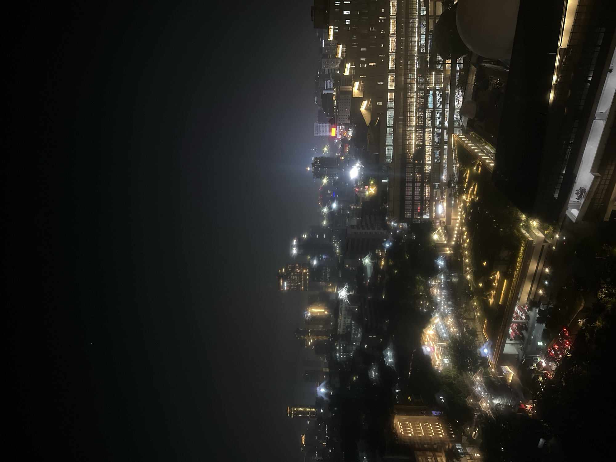 An aerial, angled, nighttime view shows the Central Jakarta, Indonesia, cityscape. Illuminated buildings rise from a dark foreground. The city is interspersed with lights, including rows of street lamps. Cars are visible on the streets. The sky is dark. The scene is hazy or misty.