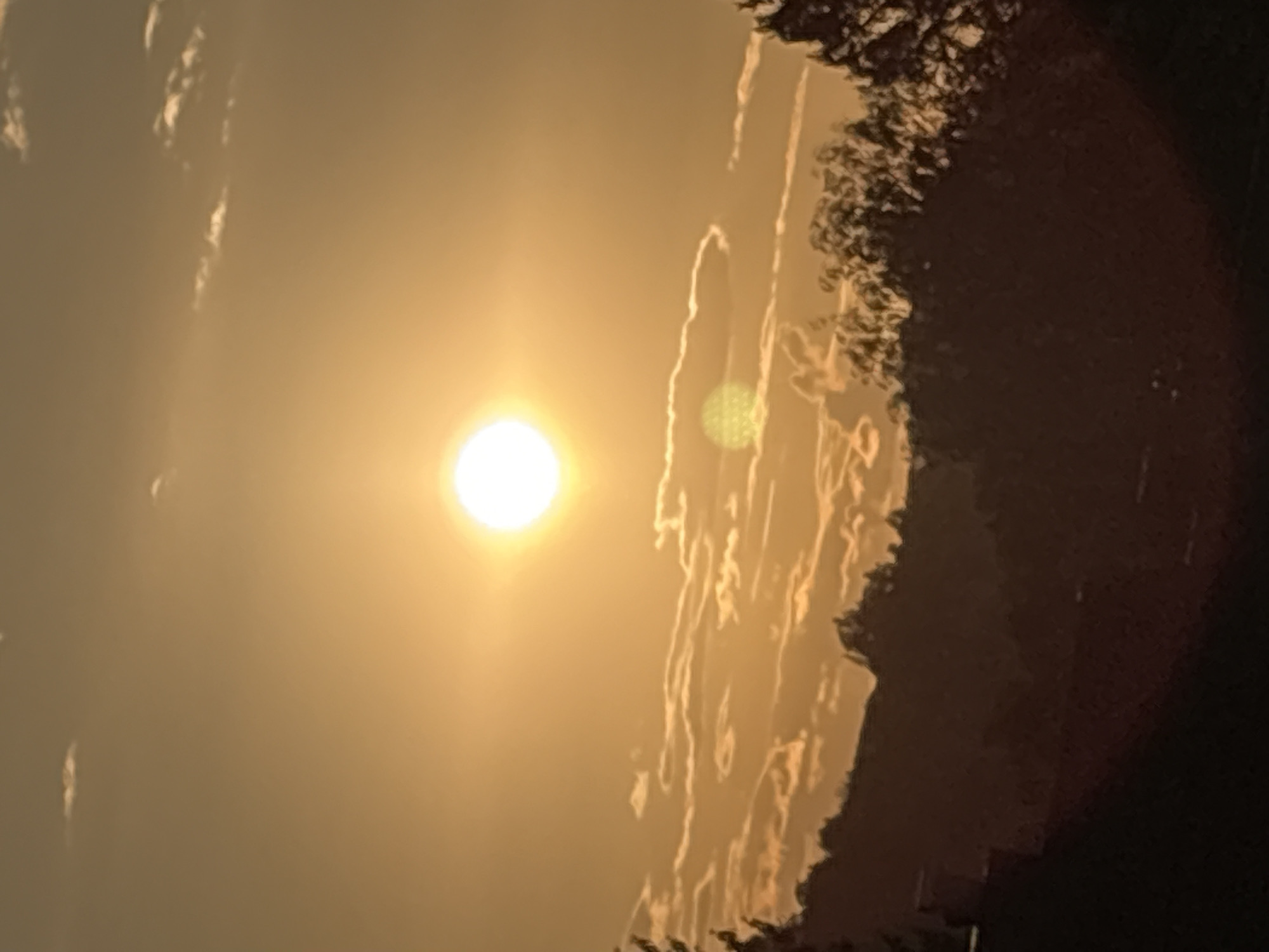 This is a photograph of a sunset over the city of Osterode am Harz, Germany. The sun is a bright, glowing orb in the center of the frame, casting a warm, orange light across the sky. Wispy clouds are scattered across the sky, illuminated by the setting sun. In the foreground, silhouetted against the bright sky, are the dark shapes of trees and hills, suggesting a natural landscape on the outskirts of the city. The overall impression is one of a peaceful and serene evening.