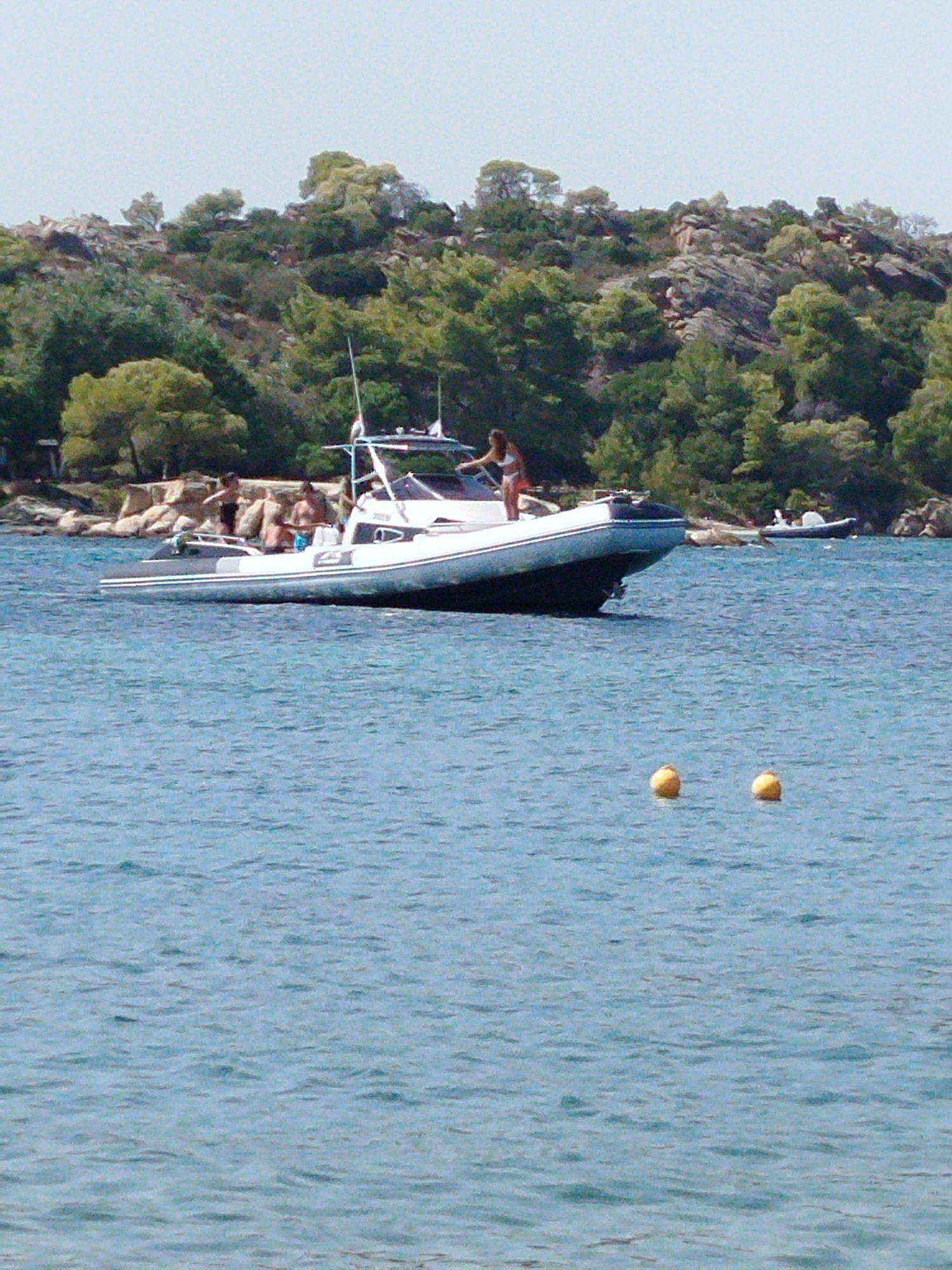 The image depicts a sunny day at a coastal location, likely in Sithonia Municipal Unit, Greece, given the typical landscape of rocky shores with lush green vegetation and clear blue water. In the foreground, the calm blue water shows ripples and two yellow buoys floating on the surface. A white and grey rigid inflatable boat (RIB) is prominently featured, moving through the water. On board the RIB, there are several people. One individual, a woman in a white bikini, is standing towards the front of the boat, appearing to be gesturing or posing. Another person, a man in black shorts, is standing near the rear of the boat. Two other individuals, also appearing to be men, are seated or standing further back. In the midground, a tree-covered, rocky hillside rises from the water's edge. The vegetation is dense and varied, with shades of green indicating a healthy ecosystem. Further in the background, the sky is a pale blue and clear, suggesting good weather. A small, white boat is visible near the shoreline in the distance. The overall scene conveys a leisurely atmosphere, typical of a summer day at a Greek coastal resort.