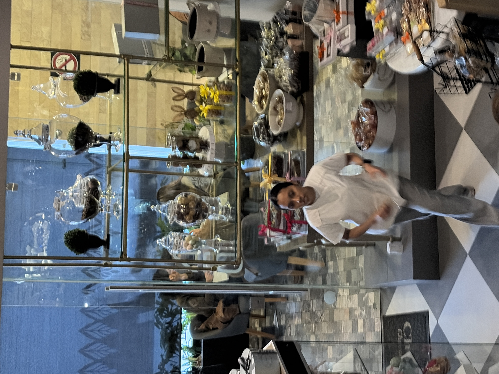 The image presents an interior view of a commercial establishment, likely a confectionery shop or cafe, situated in Puebla, Mexico. The perspective is elevated, looking down into the space.
In the immediate foreground, a person is walking towards the viewer, positioned centrally and slightly to the right, exhibiting motion blur. This individual is dressed in a white short-sleeved V-neck top and light gray trousers, indicative of a uniform. The flooring in the foreground consists of a checkerboard pattern of alternating light and dark gray square tiles, transitioning to rectangular gray tiles further back.
A central, multi-tiered display unit constructed of metal and glass shelving dominates the mid-ground. The upper shelves feature multiple large, clear glass apothecary jars containing dark granular material, flanked by small potted faux plants. A red and white "No Smoking" sign is affixed to the right side of the upper shelving. The middle shelves hold additional glass jars, some filled with colorful spherical candies, alongside two decorative rabbit figurines adorned with yellow bows. Lower shelves and counters showcase various bowls and containers filled with assorted confectionery items, including individually wrapped chocolates, pastries, and packaged gift boxes with red ribbons. A partially visible white round table on the lower right displays more packaged treats and a black wire basket.
Beyond a clear glass partition in the background, a seating area is visible. At least three individuals are seated at light-colored tables on gray upholstered chairs with wooden legs. One person on the left is seated, with a brown bag or item on an adjacent chair. The distant background behind the seating area features a dark blue wall or blind with a repetitive light-colored leaf motif, and a lighter-colored wall section to the upper right. A dark floor mat with obscured light-colored text, possibly "CHOC," is visible near a glass door on the far left. The environment is uniformly illuminated by interior lighting.